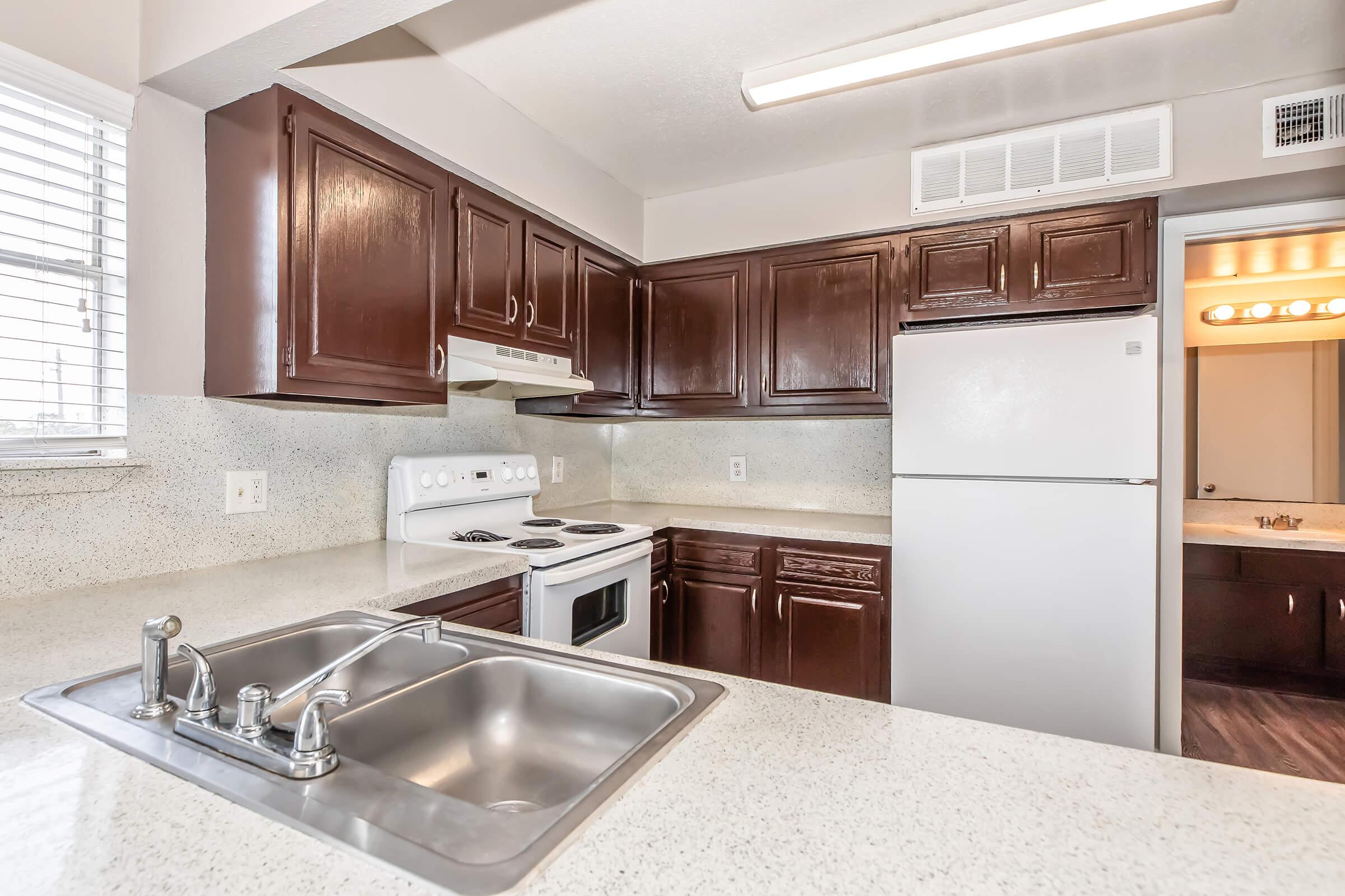Modern kitchen featuring dark brown wooden cabinets, a white stove and oven, a stainless steel sink with a double basin, and a white refrigerator. Light-colored countertops and a window provide natural light, while a doorway leads to another room, hinting at a spacious layout.