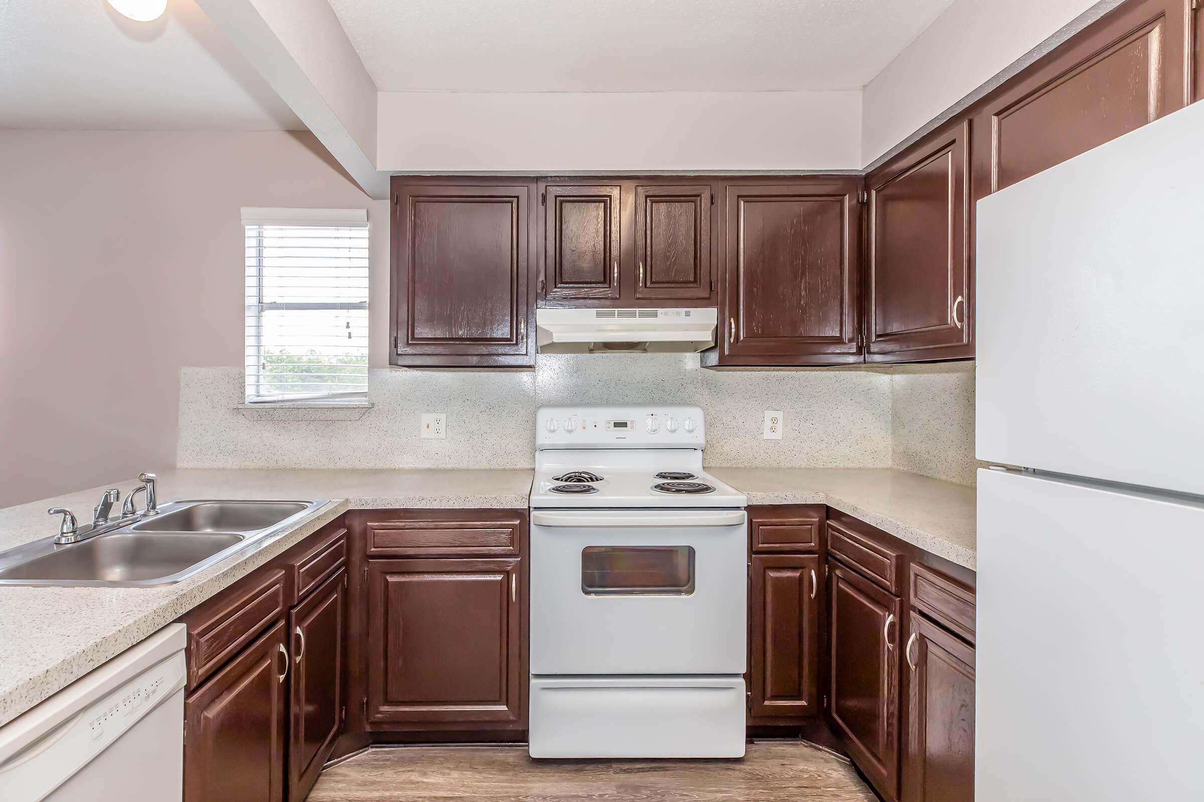 A modern kitchen featuring dark wood cabinets, a white stove and hood, a white refrigerator, and a double sink. Natural light enters through a window above the sink, illuminating the beige countertops and the flooring. The kitchen has a clean and functional layout suitable for cooking and meal preparation.