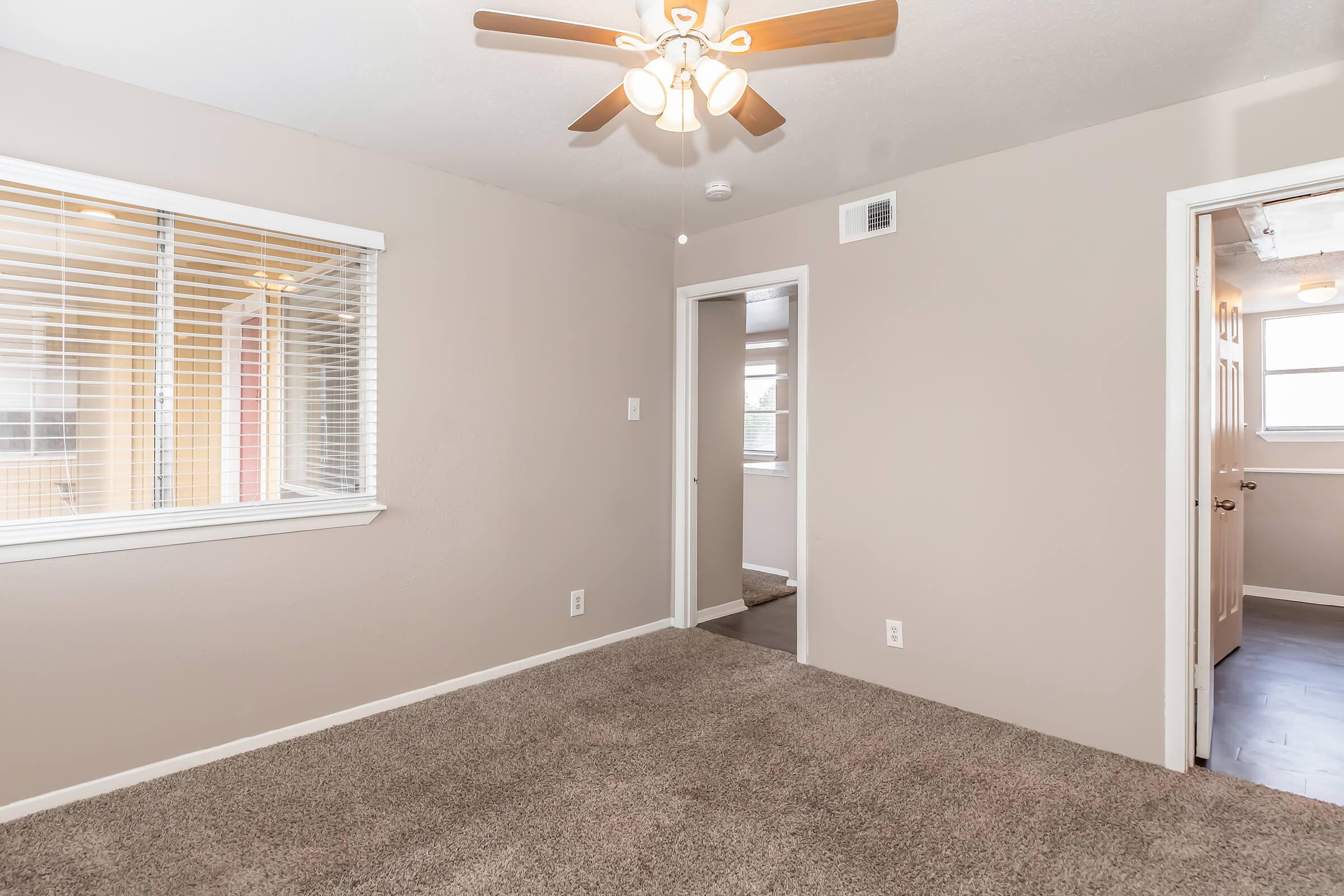 A well-lit room featuring beige walls and carpet, with a ceiling fan. One wall has a window with blinds, and there's a doorway leading to another space. The room is empty, emphasizing its spaciousness and neutral decor.