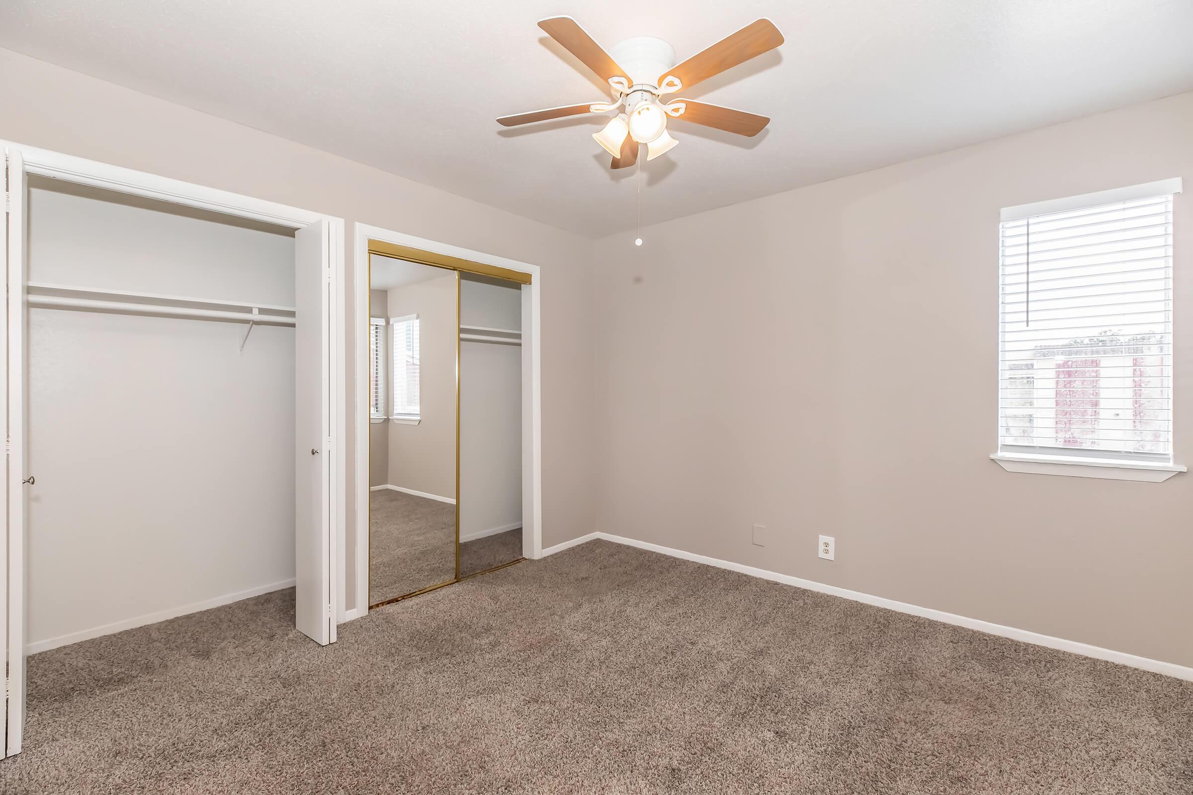 A spacious, empty bedroom with light beige walls and carpet. It features a ceiling fan, a large mirrored closet with sliding doors, and two windows with white blinds allowing natural light to enter. The room is well-lit and has a clean, inviting atmosphere.