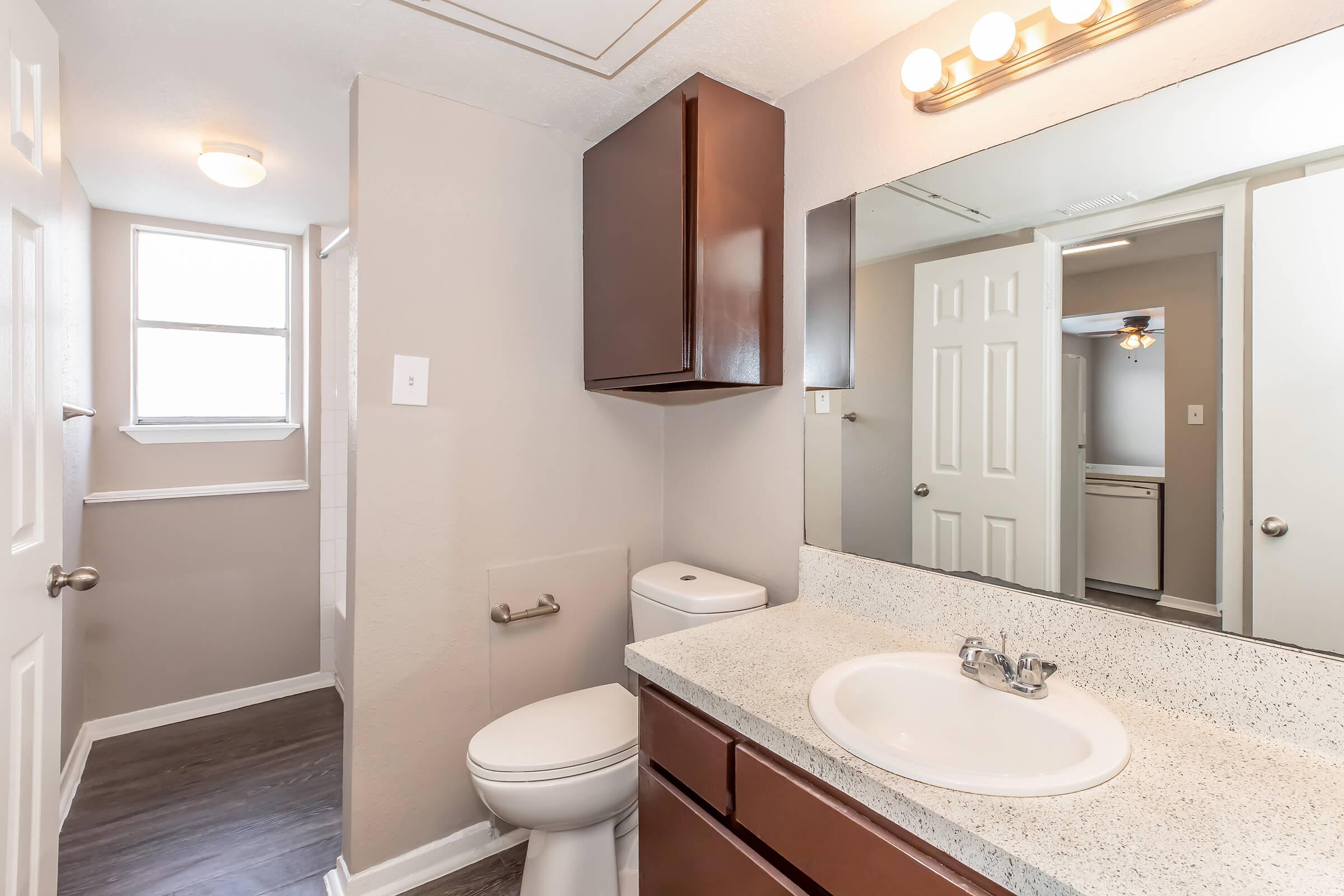 A modern bathroom featuring a white sink with a granite countertop, a wooden cabinet above the sink, a toilet, and light fixtures. The walls are painted in a neutral tone, and there is a mirror reflecting the room. A doorway leads to a hallway, with natural light coming from a nearby window.
