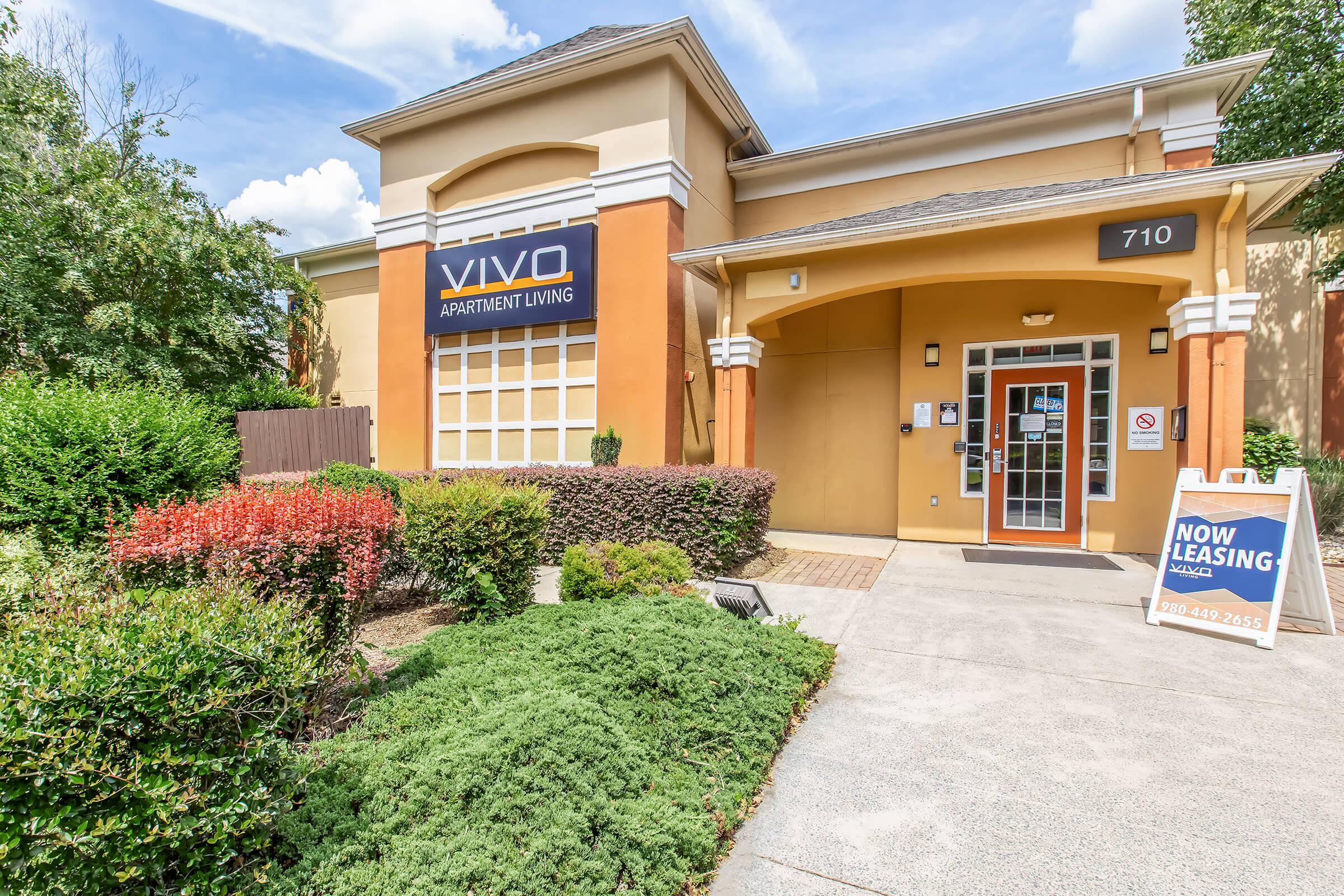 Exterior view of Vivo Apartment Living building with a welcoming entrance. The building features a combination of orange and beige colors, surrounded by well-maintained greenery. A sign displays "NOW LEASING" on an A-frame board near the entrance. Clear blue skies above enhance the inviting atmosphere.