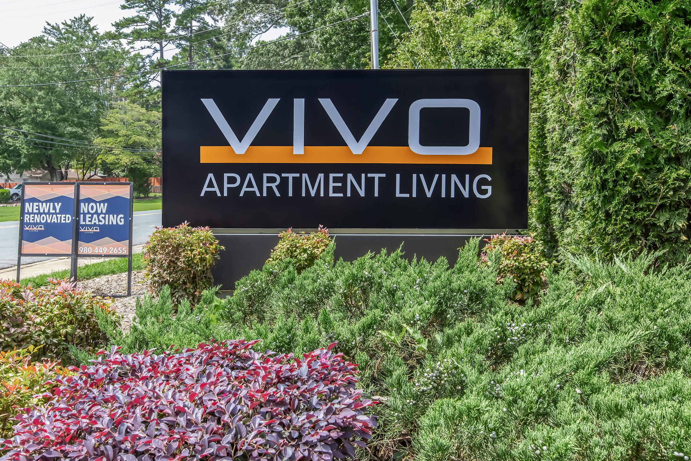 Sign for Vivo Apartment Living, featuring the name "VIVO" prominently in large letters. Surrounding the sign are well-maintained shrubs and colorful plants. To the left, there are additional signs indicating "Newly Renovated" and "Now Leasing" with a phone number displayed.