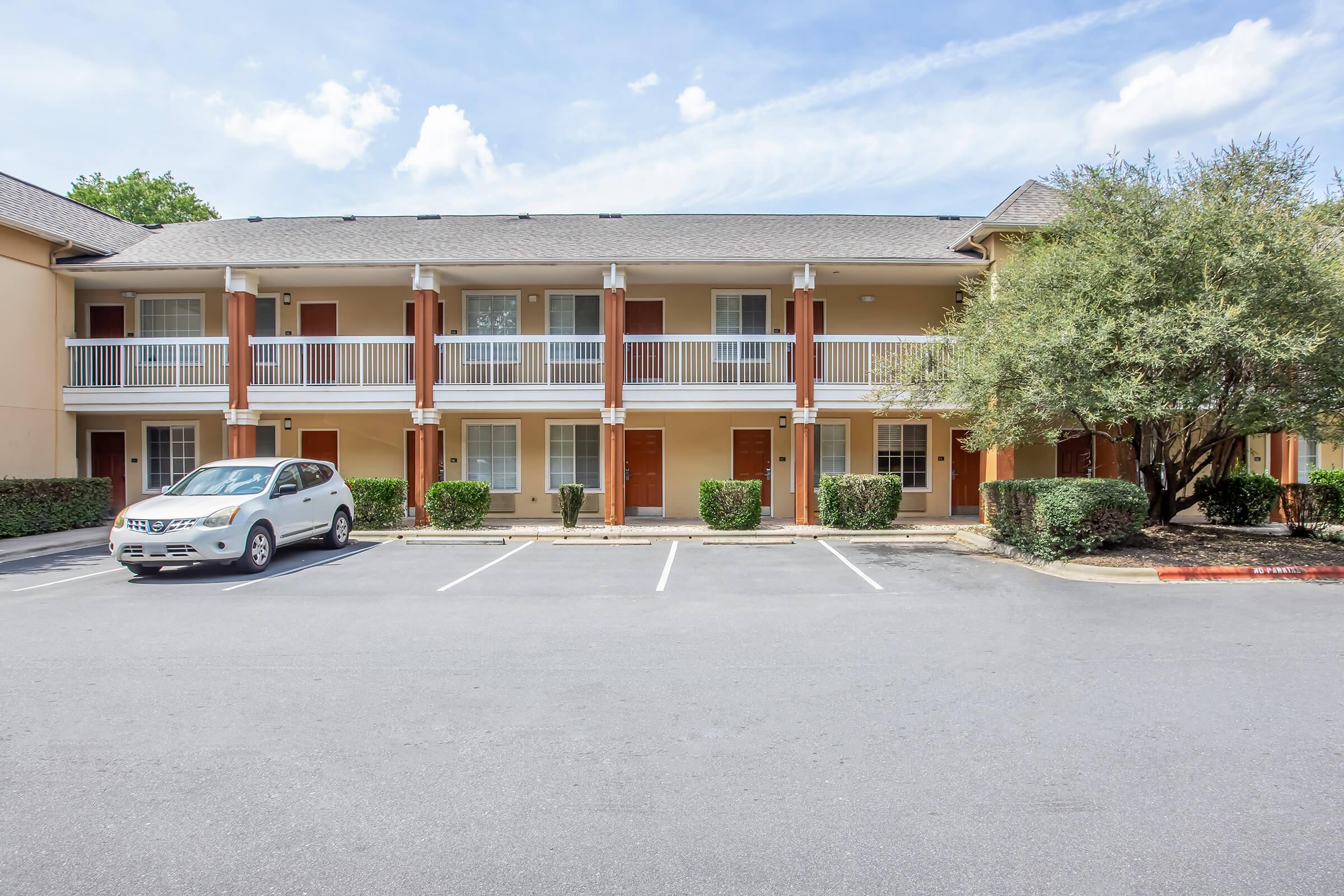 A view of a two-story hotel or motel building featuring multiple rooms with balconies. Each room has a door, and the exterior is adorned with landscaping. A silver SUV is parked in front of the building, and the sky is partly cloudy. The parking area is spacious with marked spots.