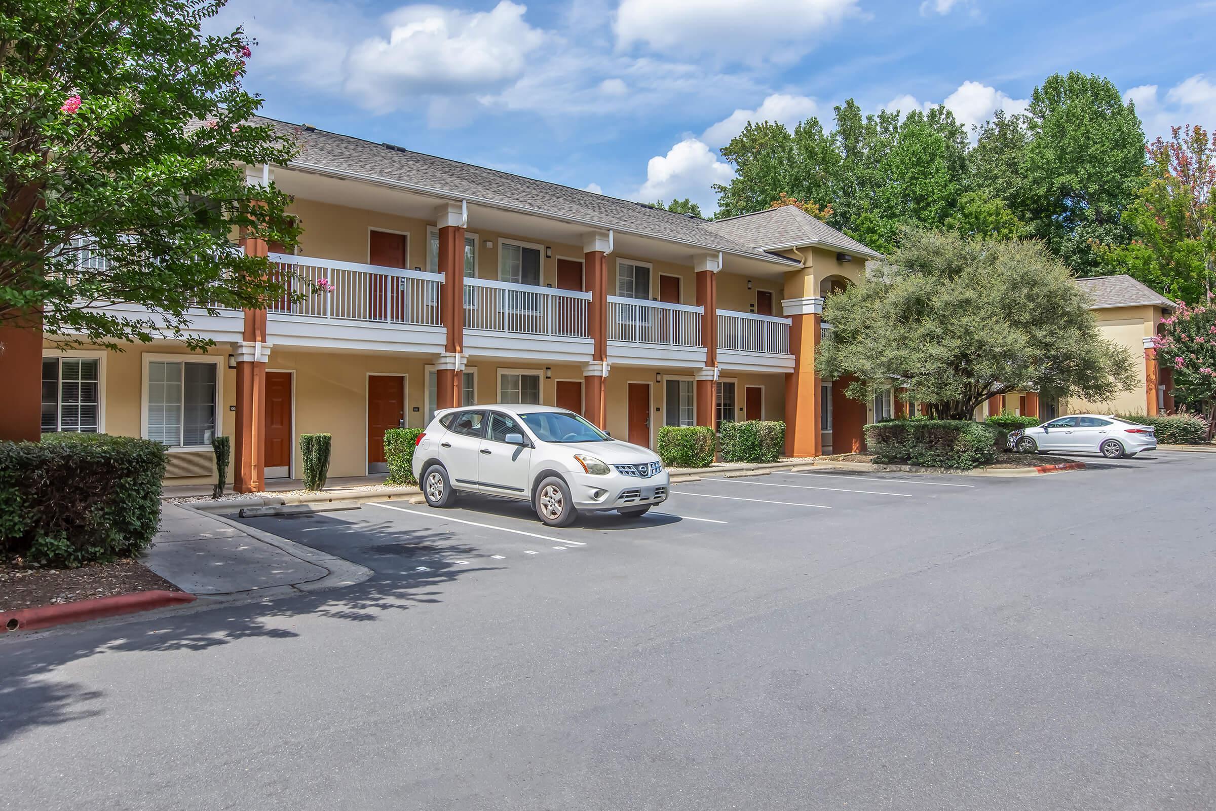 A view of a two-story building with multiple individual units, featuring balconies and orange accents. There are two parked cars in the foreground, surrounded by landscaped shrubbery and trees under a partly cloudy sky. The setting appears to be a residential or motel area.
