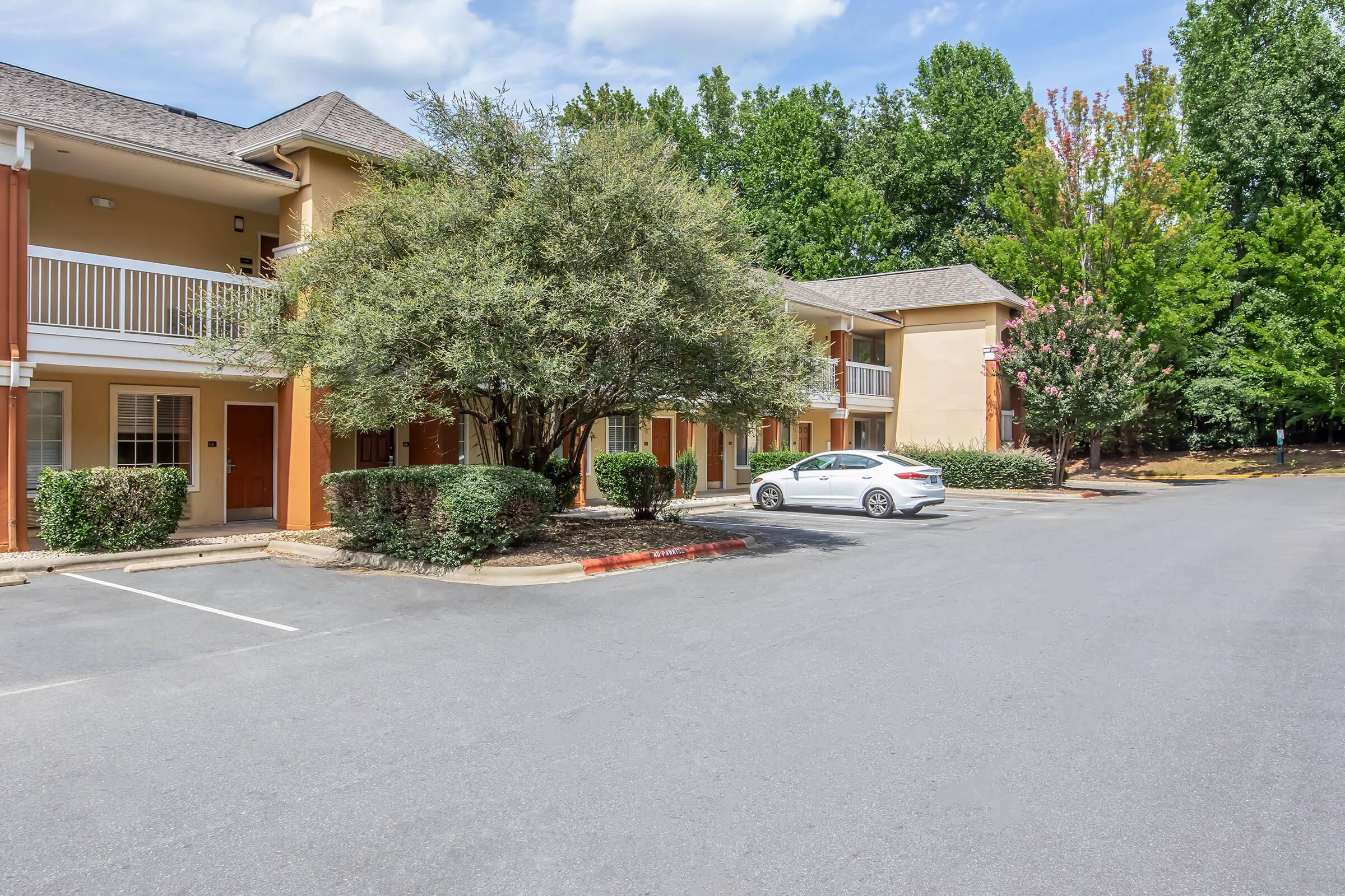 A view of a quiet hotel exterior featuring orange and beige buildings, surrounded by green trees and shrubbery. A white car is parked in the lot, and the scene is set under a blue sky with scattered clouds. The area appears well-maintained and inviting for guests.