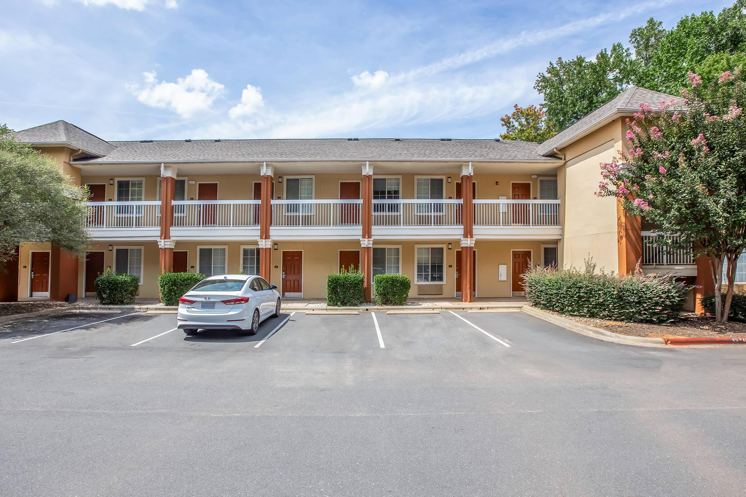 A two-story building with multiple guestroom balconies, featuring a light-colored exterior and orange accents. A white car is parked in the foreground, and neatly trimmed bushes line the sides of the entrance. The sky is blue with some clouds, creating a bright atmosphere.