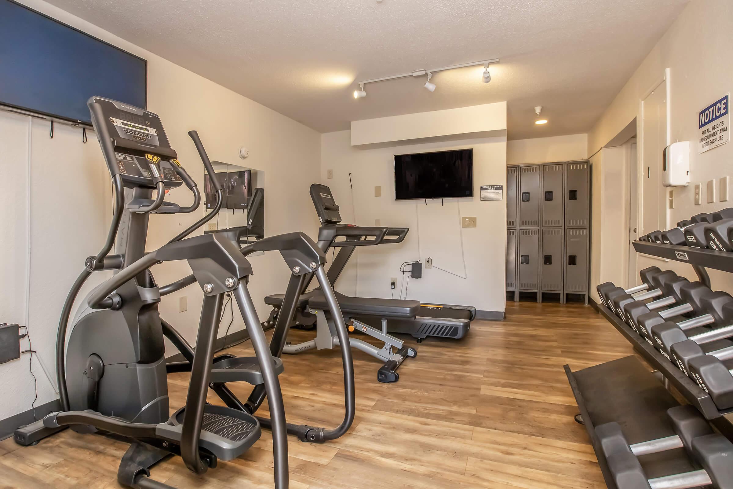 A fitness room featuring various exercise equipment, including treadmills and elliptical machines, with a TV on the wall. There are also weights on a rack and lockers in the background, all set in a light-colored environment with wooden flooring.