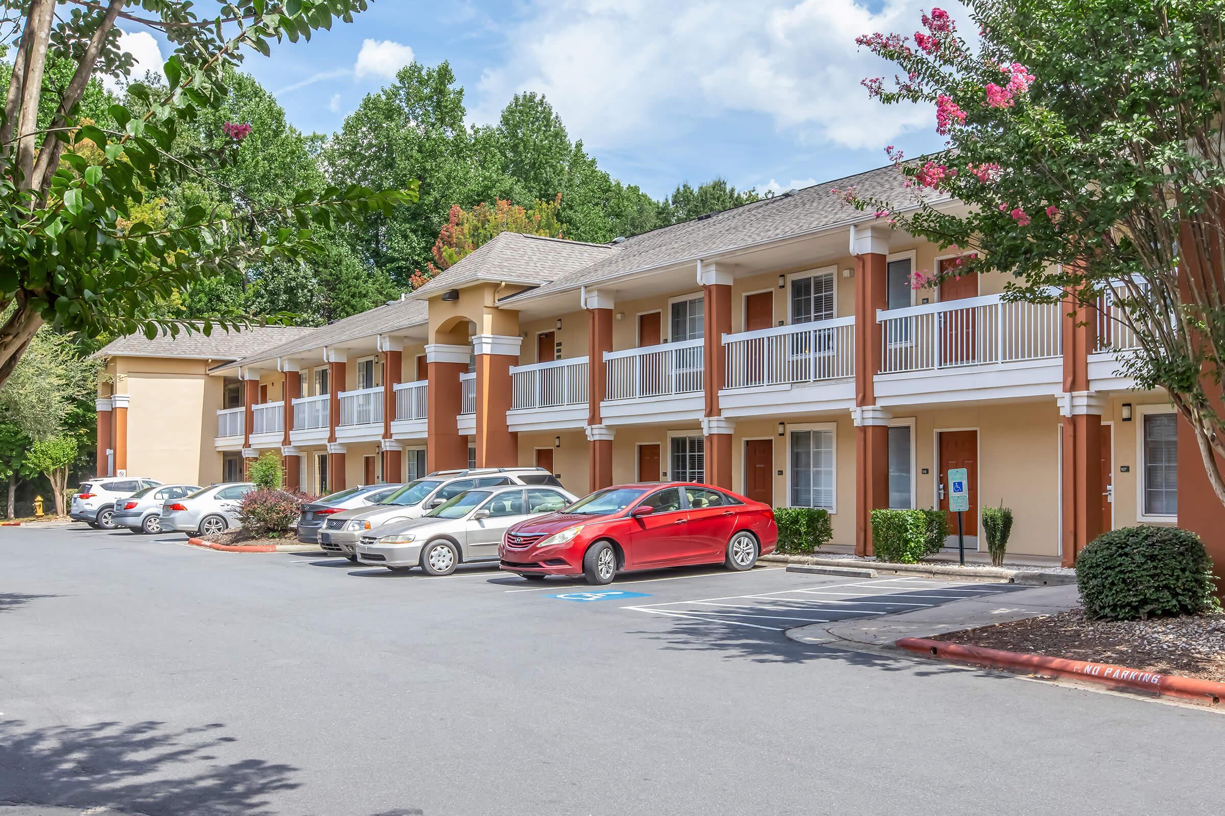 Exterior view of a multi-story hotel with a beige and orange facade, featuring balconies. Several parked cars are visible in the lot, including a red sedan. The surroundings include green trees and landscaping, with a blue sky and scattered clouds in the background.