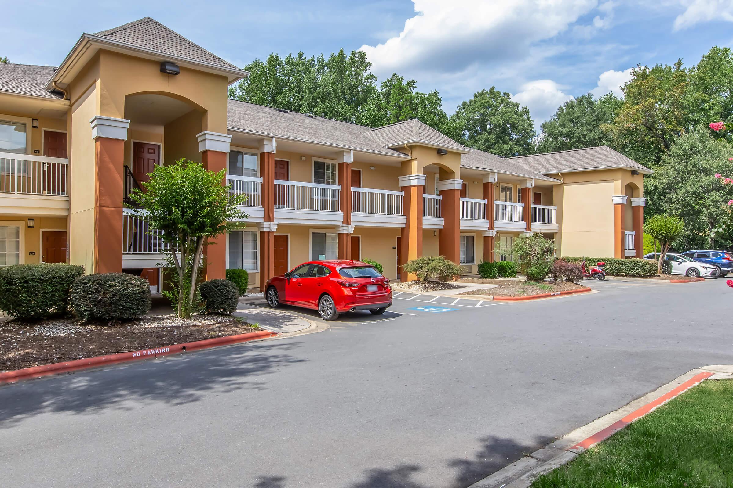 Exterior view of a multi-story building featuring residential apartments. A red car is parked in front, surrounded by neatly trimmed shrubs and small trees. The sky is partly cloudy, and there are additional vehicles in the parking area, illustrating a well-maintained community.