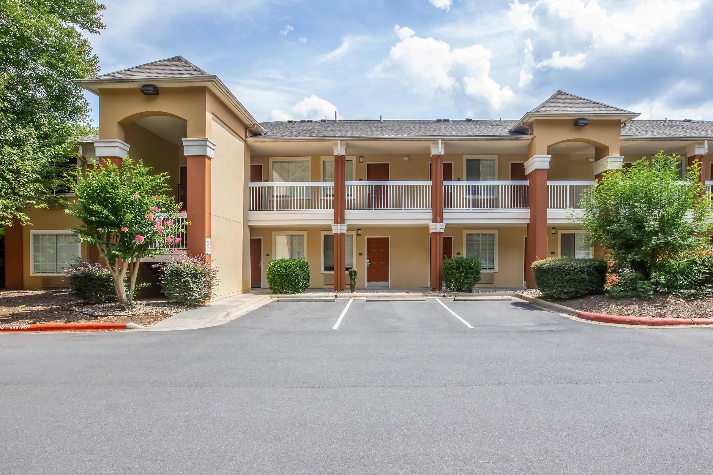 Exterior view of a two-story hotel building featuring multiple entrances, balconies with railings, and landscaped greenery. The parking area is visible in front, with well-maintained shrubs and trees nearby, under a partly cloudy sky. The architecture is modern and inviting.
