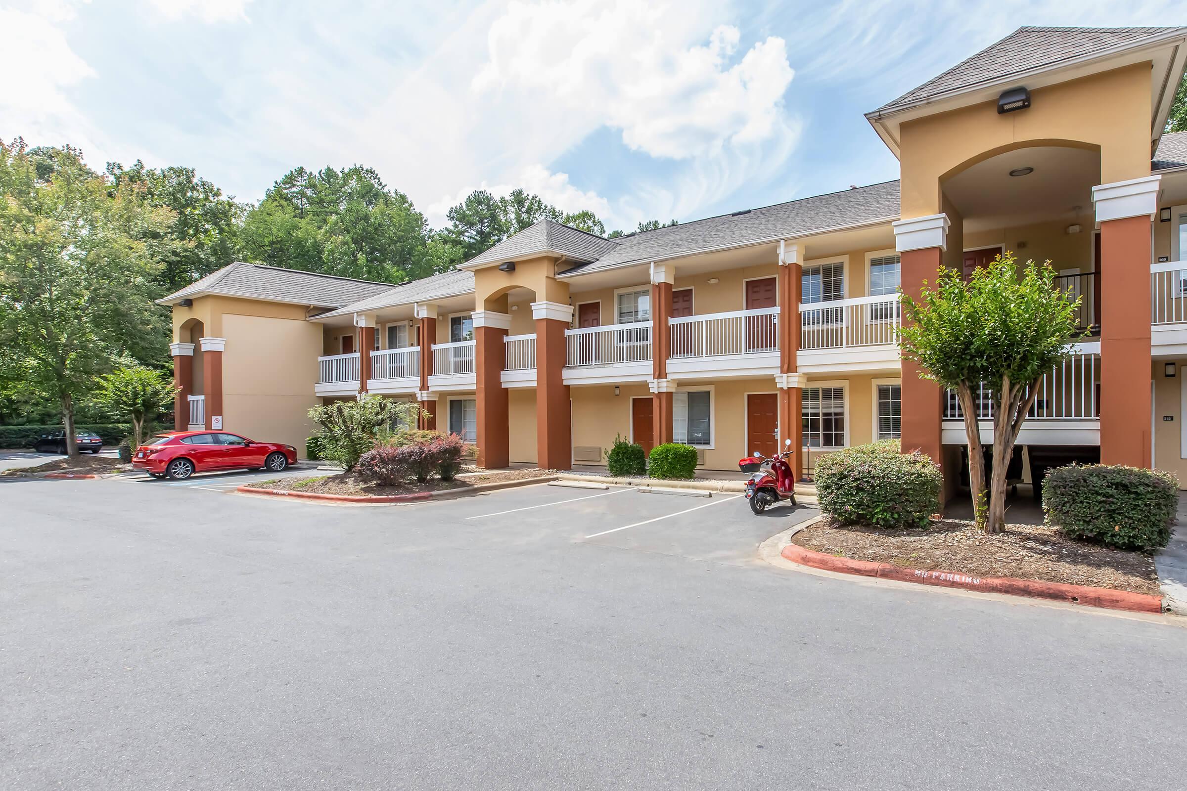 A view of a multi-story apartment building with a light orange exterior, featuring balconies on the upper floor. The parking area includes a red car, well-maintained shrubs, and trees in the background under a partly cloudy sky.