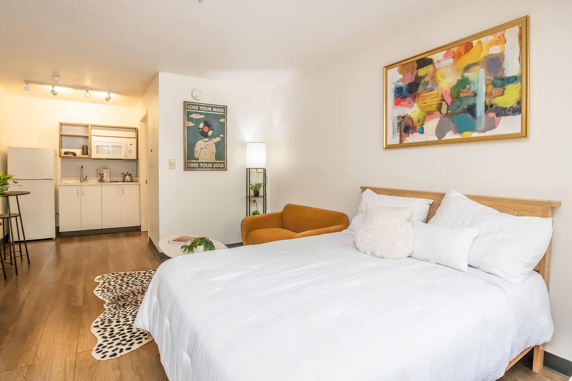 A modern studio apartment featuring a cozy bed with white bedding, a pair of decorative pillows, an orange armchair, and a small table. The kitchen area is partially visible with light cabinets and appliances. A colorful abstract painting hangs on the wall, and a leopard-print rug adds a stylish touch to the floor.