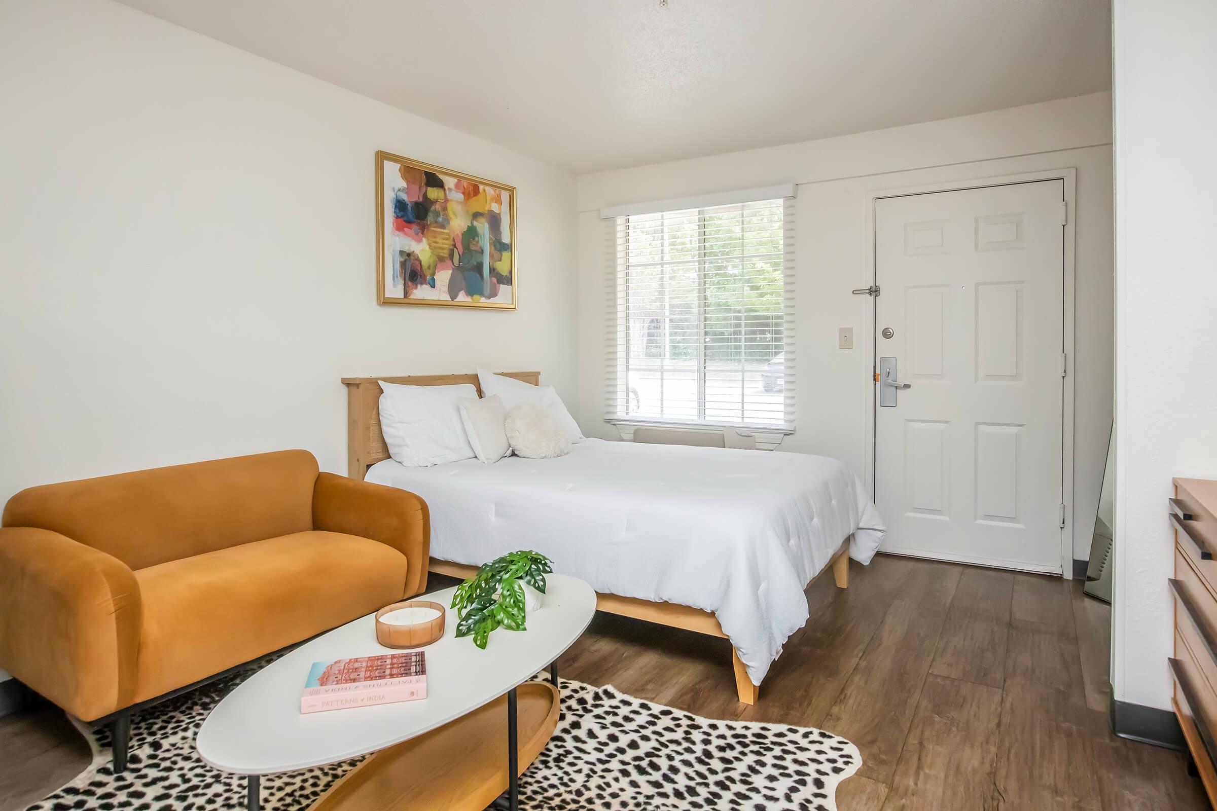 A cozy room featuring a double bed with white bedding, a stylish orange couch, a round coffee table with a book and a plant, and a door with a window. The walls are painted white, and there is a vibrant piece of artwork above the bed. Natural light spills through the window, enhancing the inviting atmosphere.
