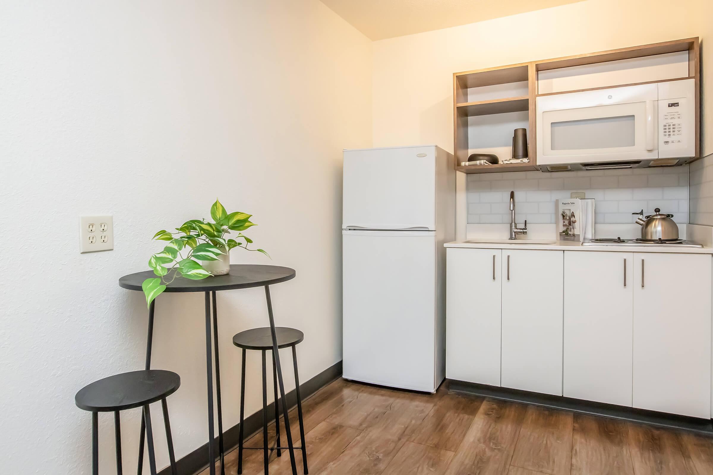 A small, modern kitchen featuring a compact dining table with two stools, a white refrigerator, a microwave, and a stovetop. There is a kettle on the countertop and a small plant on the table, with light-colored walls and dark wood flooring, creating a cozy, minimalist space.