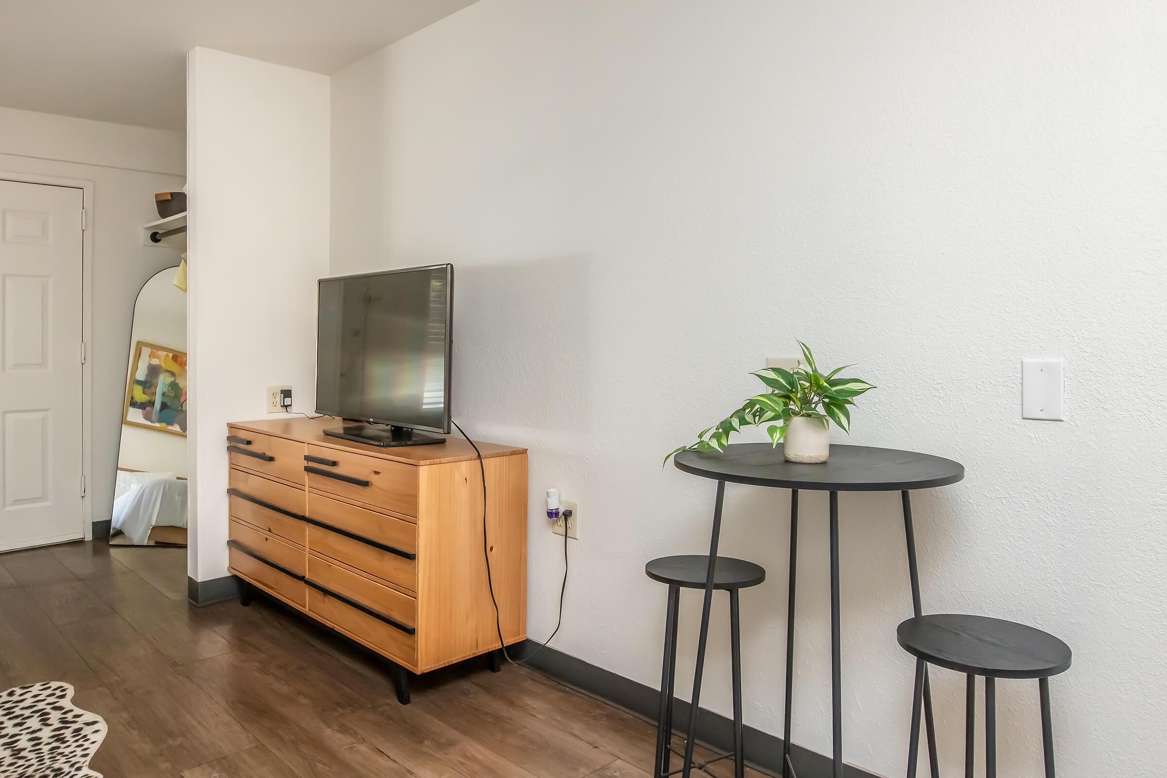 A small, modern living room featuring a television on a wooden media console, with a decorative plant on a round black table. Two black stools are positioned next to the table. The walls are painted white, and there is a glimpse of a bed and a wardrobe in the background.
