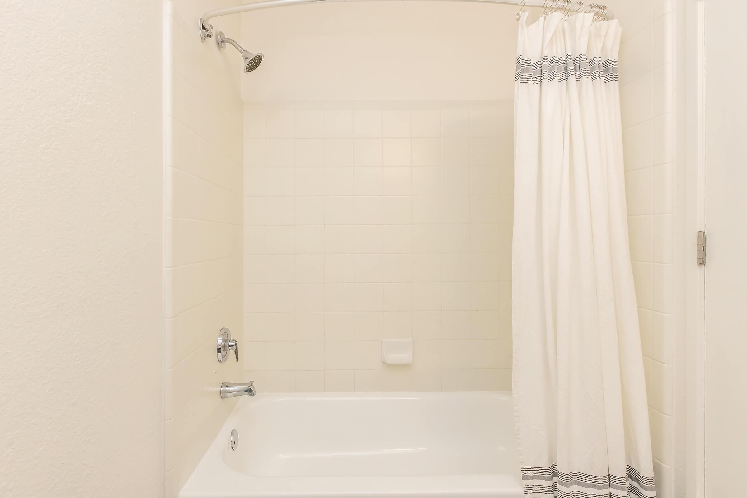 A clean, minimalist bathroom with a bathtub and a showerhead. The bathtub is white, and there is a white shower curtain with gray stripes. The walls are tiled in light colors, and the overall ambiance appears bright and spacious. The scene conveys a sense of tranquility and simplicity.