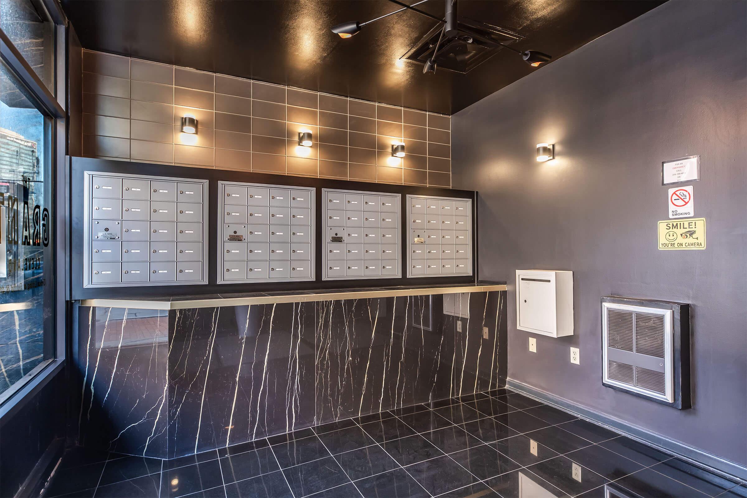 A modern mailroom featuring multiple gray mailboxes arranged on a wall. The floor is shiny black tile with a marble pattern. Soft lighting highlights the space, and there is a wall-mounted mailbox on the right side. A "Smile!" sign is visible nearby, adding a welcoming touch to the area.
