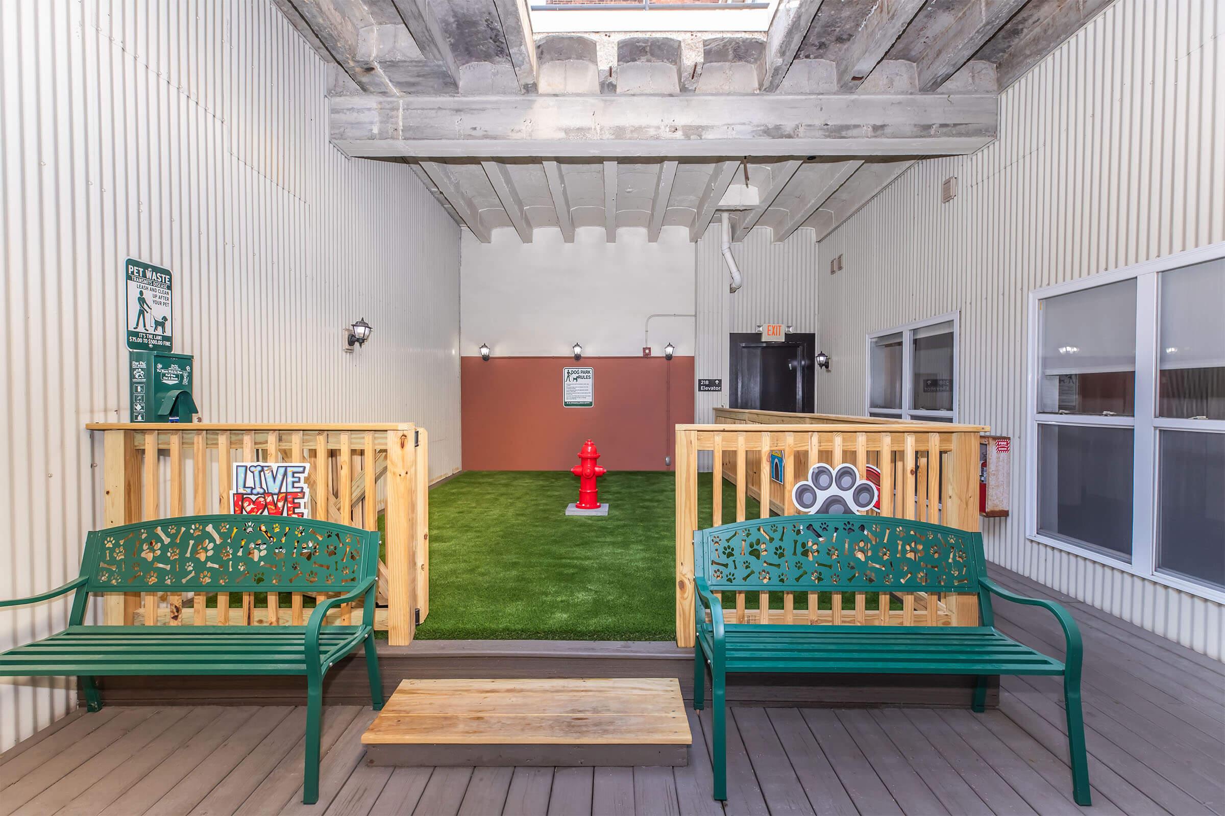 A pet-friendly outdoor space featuring green artificial grass, a red fire hydrant, and two decorative benches. The area is enclosed with wooden fencing and has adequate lighting, making it a welcoming spot for pets and their owners to enjoy.