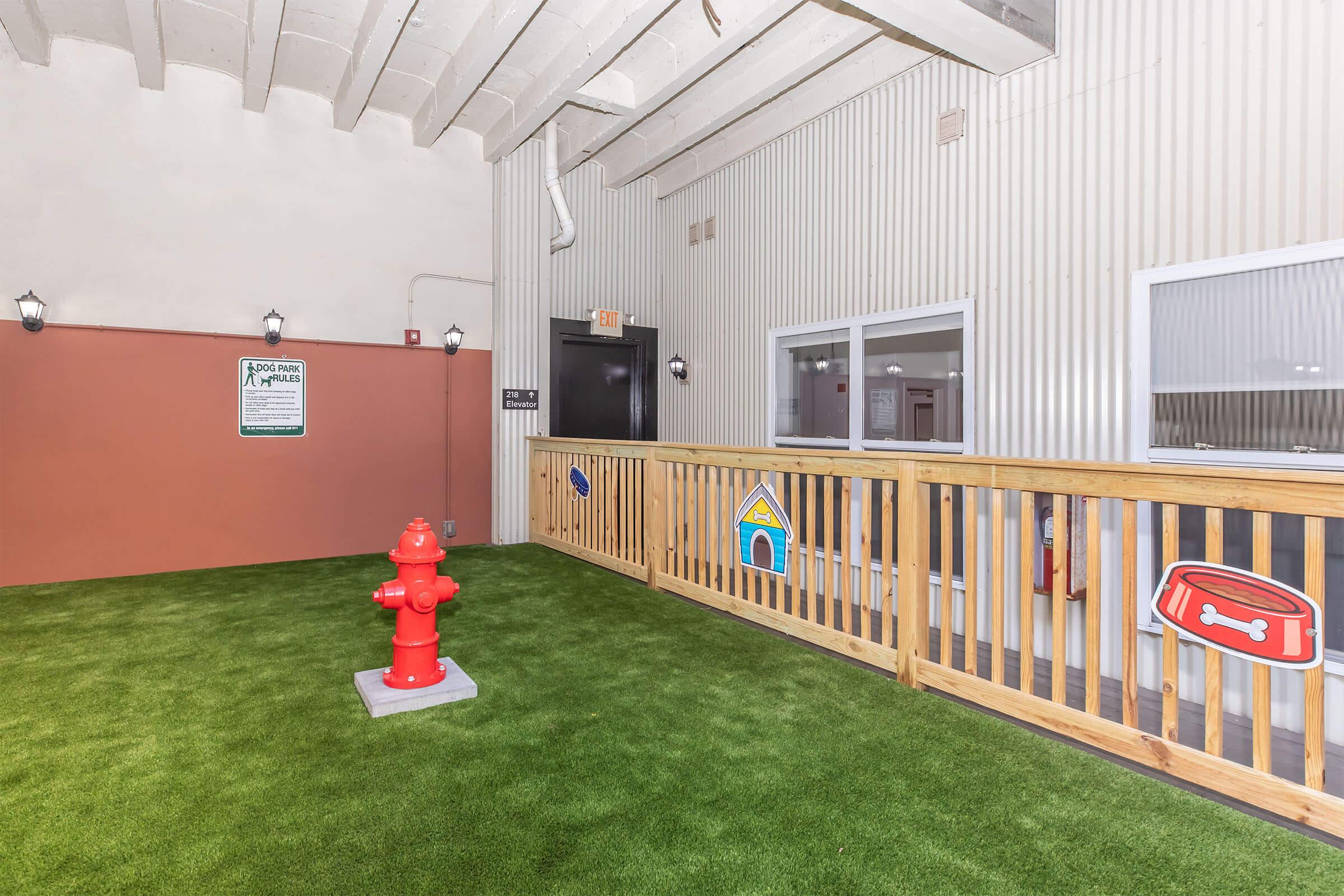 An indoor pet area featuring artificial grass, a red fire hydrant, a doghouse decoration, and a fence. There are signs on the walls related to pet policies. The space is designed for dogs, with ample room for play and potty activities.