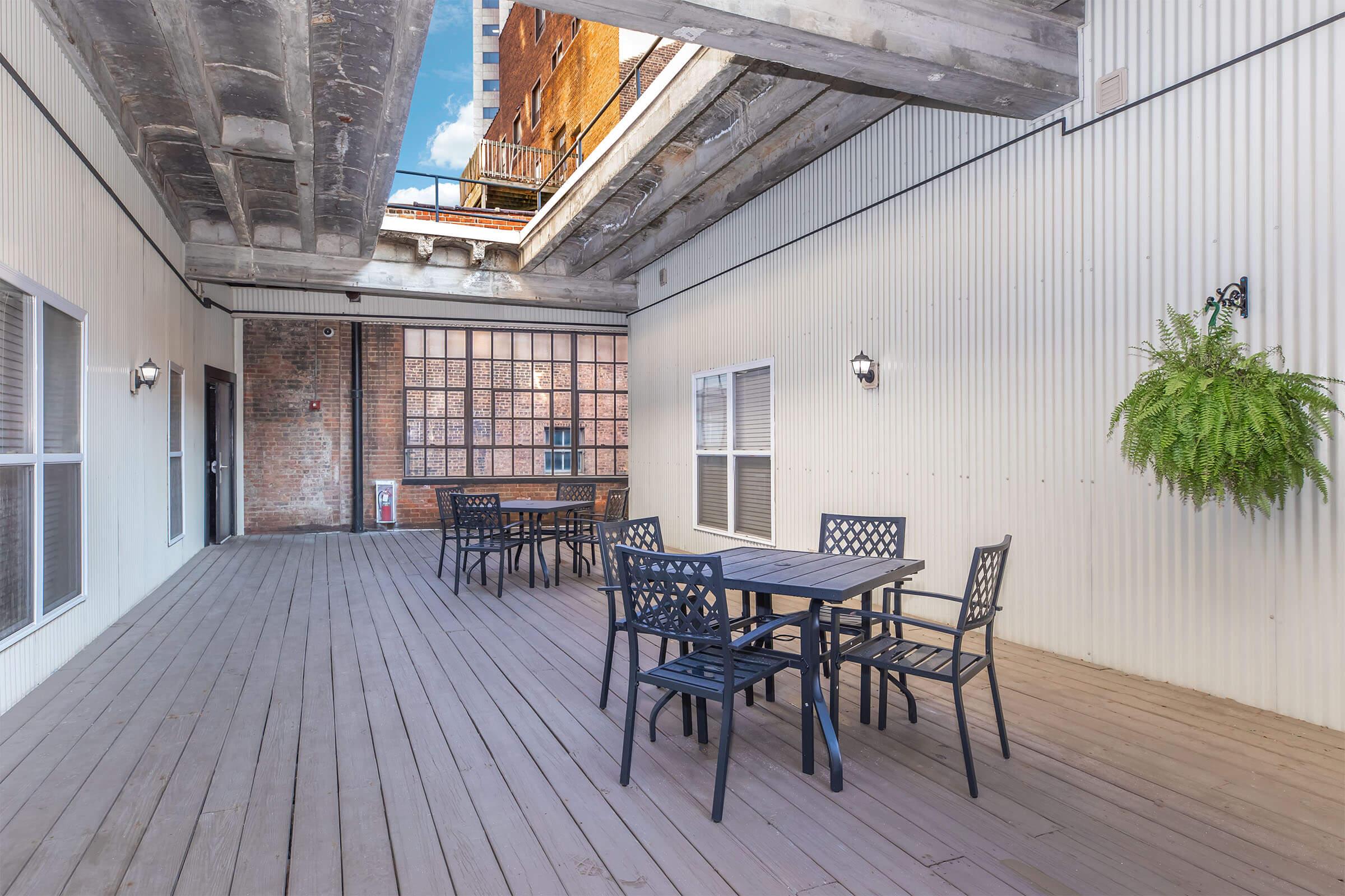 A spacious open patio area featuring a wooden floor, metal tables, and chairs. The walls have a mix of brick and metal siding, with large windows letting in natural light. Hanging ferns add greenery, and the space is illuminated by wall-mounted lights, creating a welcoming outdoor atmosphere.