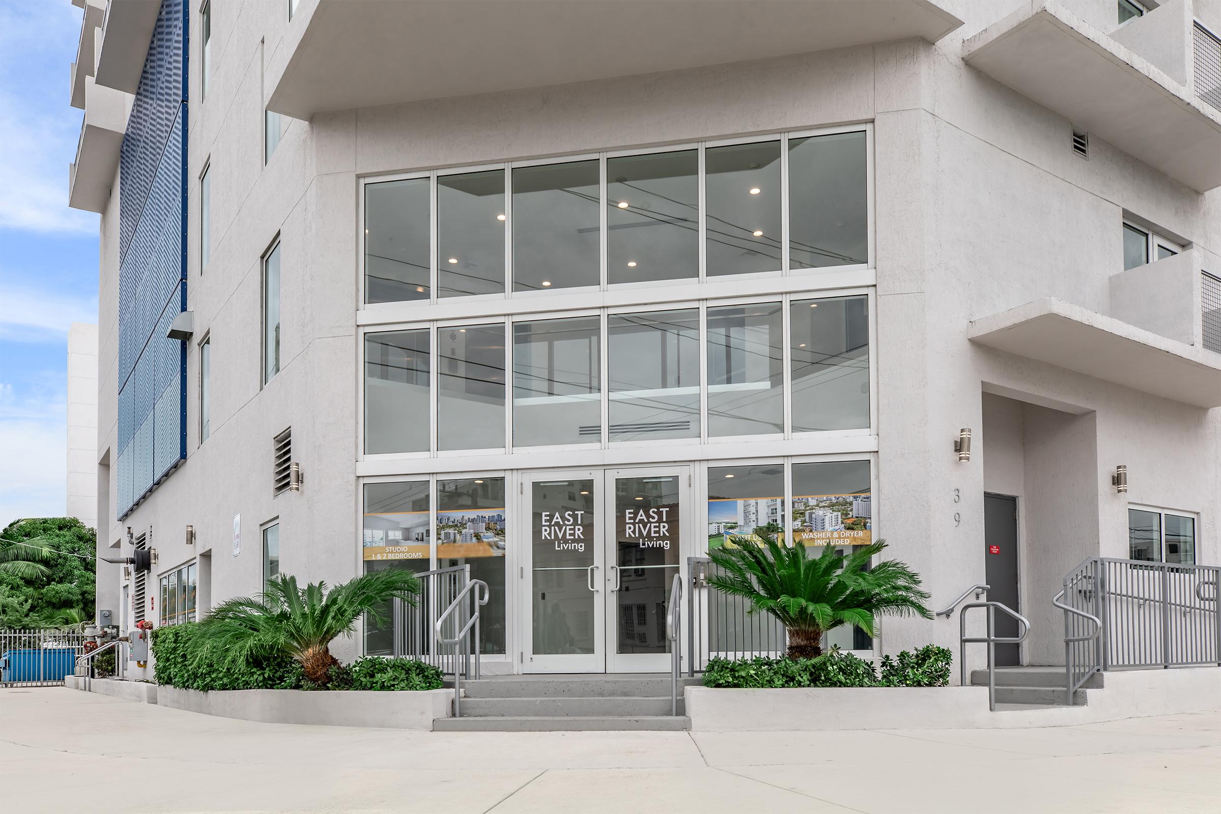 Entrance to a modern building featuring large glass doors with "EAST RIVER Living" signage. The facade is complemented by potted palm plants and a gray ramp leading to the entrance. The architecture has a clean, contemporary style, set against a bright sky and landscaped surroundings.