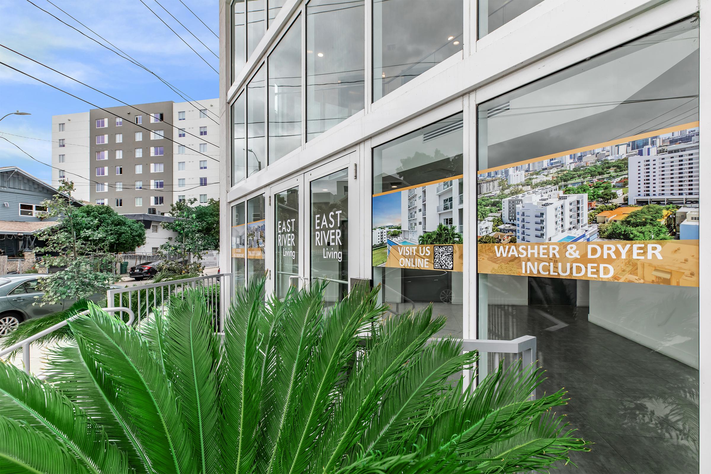 A modern apartment building with large windows showcasing promotional signs about rental options, including "East River Living" and "Washer & Dryer Included." The foreground features lush green foliage, while the background displays a multi-story building and clear sky.