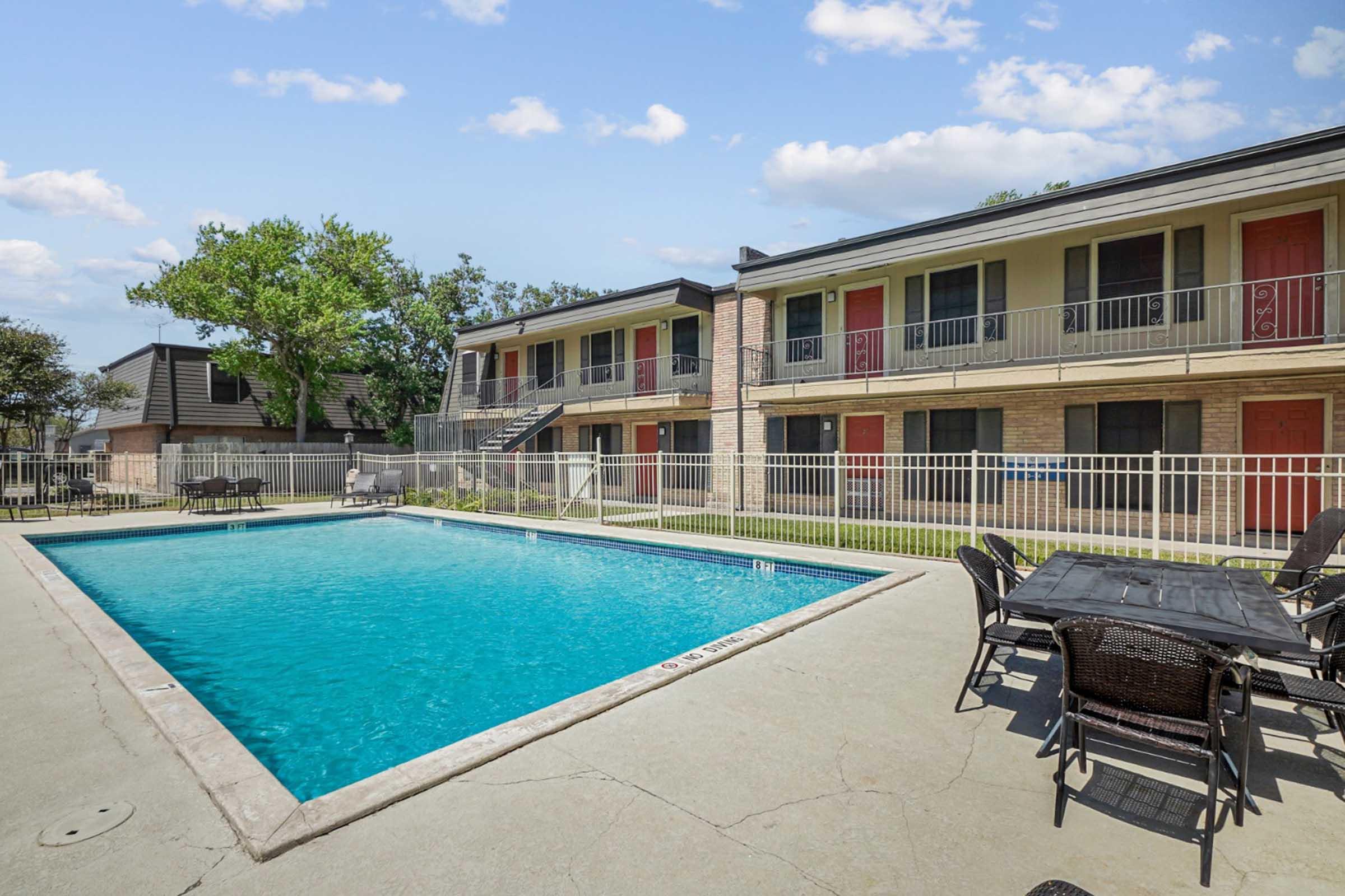 A clear blue swimming pool surrounded by a concrete deck. In the background, a two-story building with multiple apartment units, each featuring a balcony and red doors. There are outdoor tables and chairs near the pool, set against a backdrop of green trees and a partly cloudy sky.