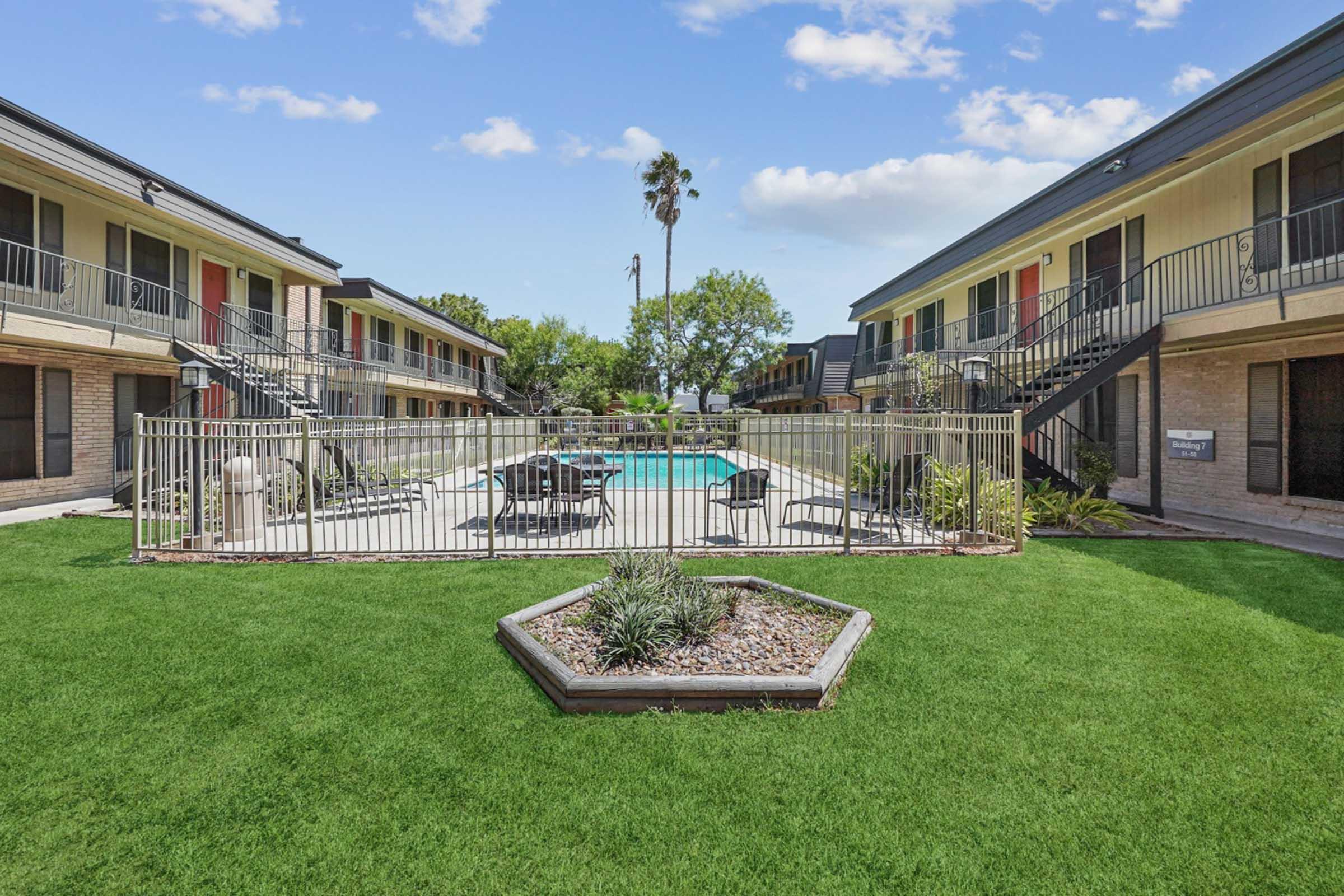 View of an apartment complex courtyard featuring a swimming pool surrounded by green grass. There are lounge chairs and tables near the pool, with palm trees visible in the background. The buildings have multiple units with balconies and colorful doors, creating a vibrant residential atmosphere.