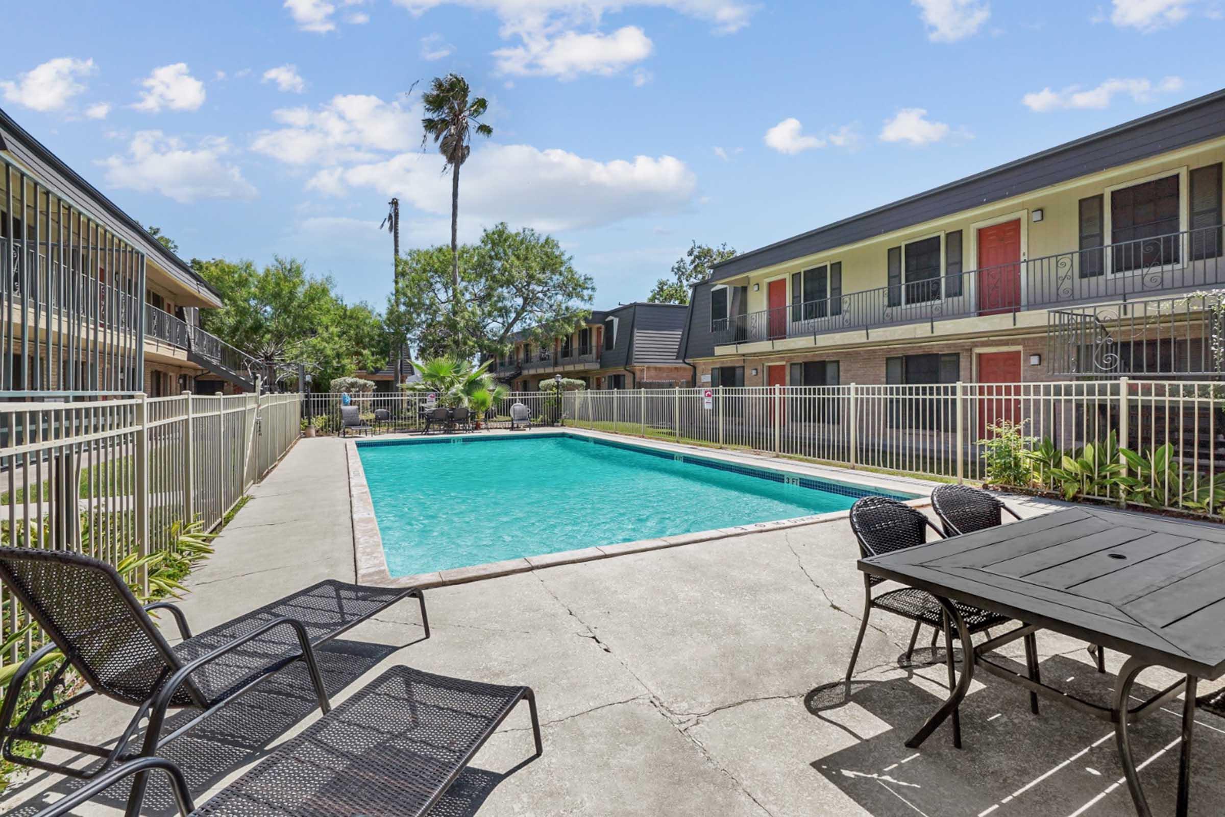 A swimming pool surrounded by a fenced area, with several lounge chairs positioned nearby. In the background, two-story apartment buildings with balconies and palm trees are visible against a clear blue sky. The scene is inviting and well-maintained, ideal for relaxation.