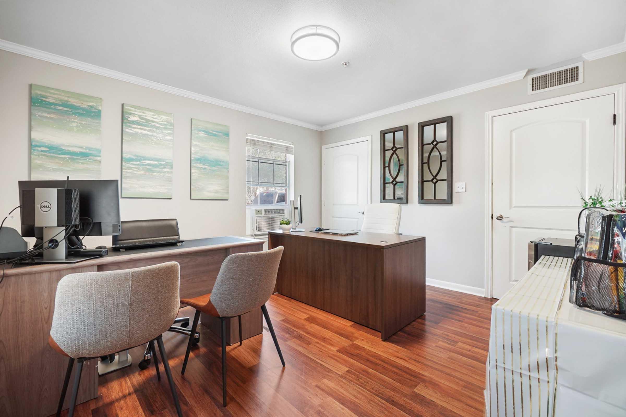 Modern office interior with wooden flooring, featuring a desk and chair, computer setup, and decorative wall art. Light colors create a bright atmosphere. A side table and additional chairs enhance the functionality of the space. Reflections in wall mirrors add depth to the room.