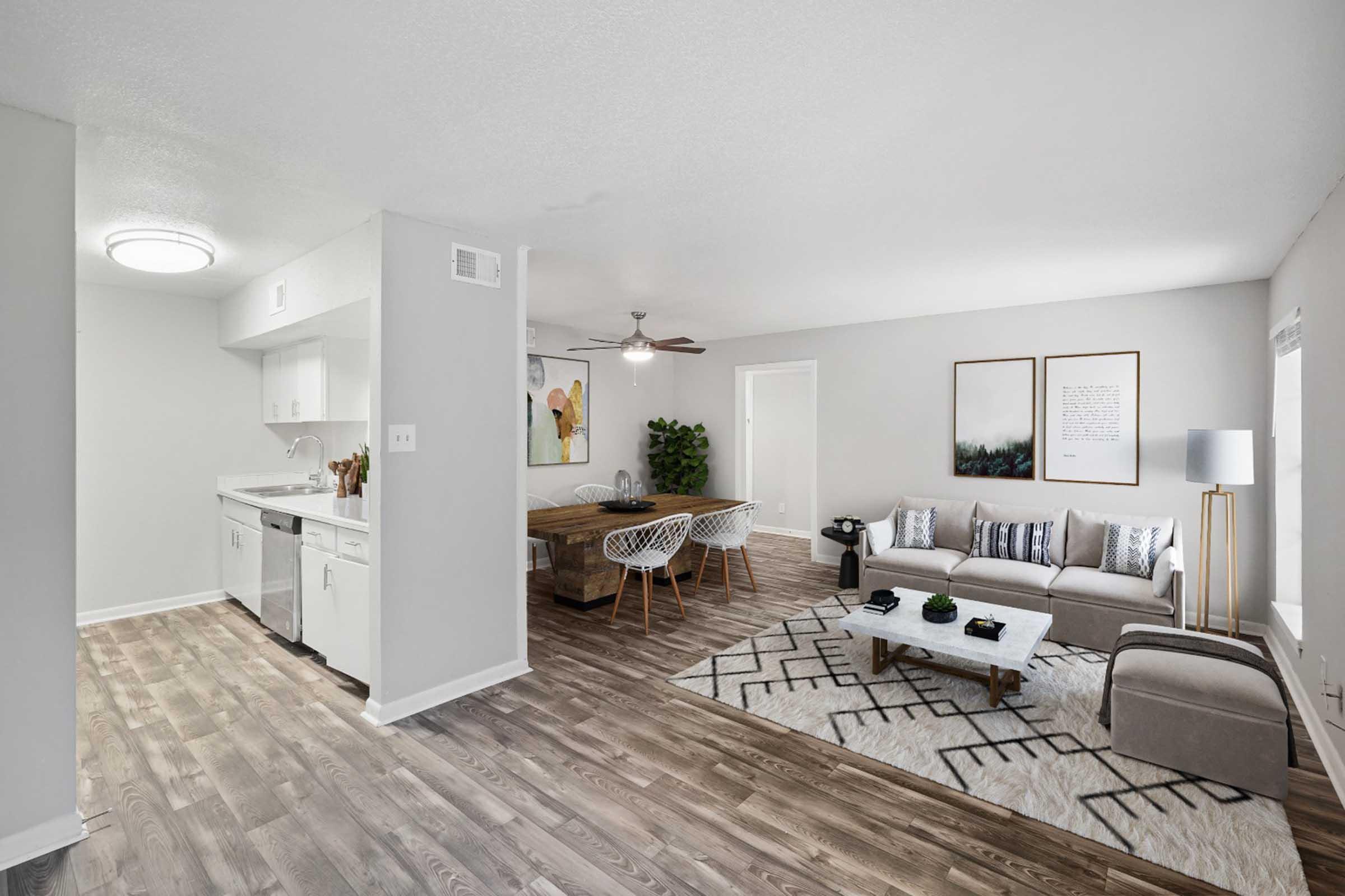 A modern open-concept living space featuring a kitchen with white cabinetry, a dining area with a wooden table and chairs, and a comfortable living room with a gray sofa, decorative pillows, and a coffee table on a patterned rug. Natural light fills the room from a window.