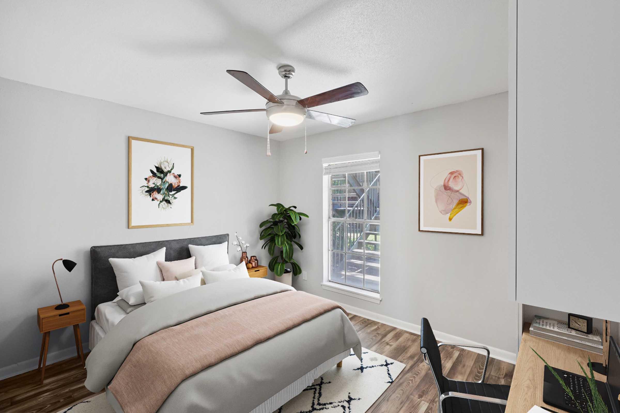 A modern bedroom featuring a gray bed with a light pink blanket, accented by white and gray pillows. There's a wooden nightstand with a black lamp, a potted plant in the corner, and large windows allowing natural light. Two framed art pieces adorn the walls, and a workspace with a chair and desk is visible.