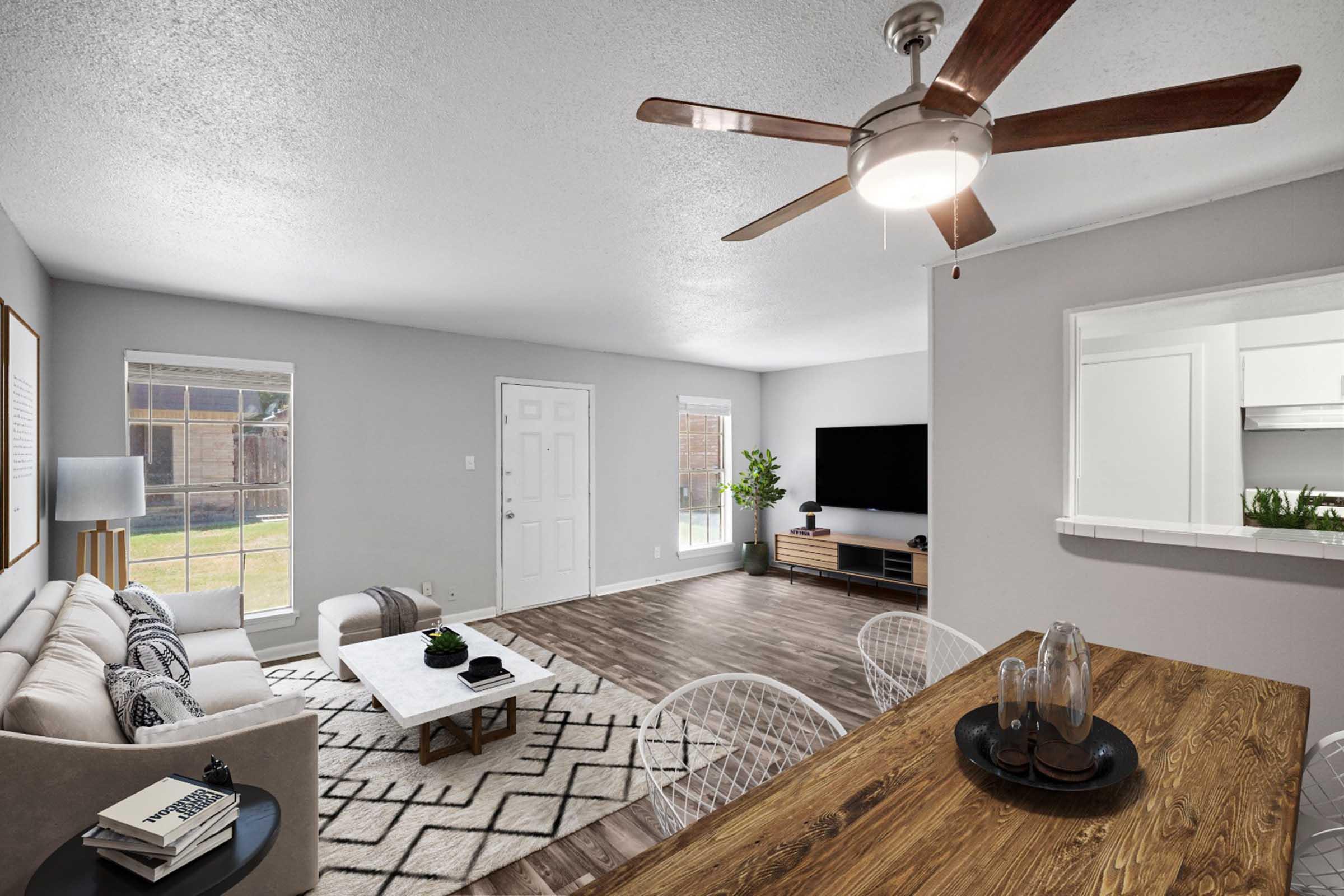 A modern living room featuring a light gray color scheme, a comfortable sofa, a stylish coffee table, and a flat-screen TV mounted on the wall. There are large windows allowing natural light, a ceiling fan, and a dining area with wooden furniture. Decorative elements include a geometric rug and potted plants.