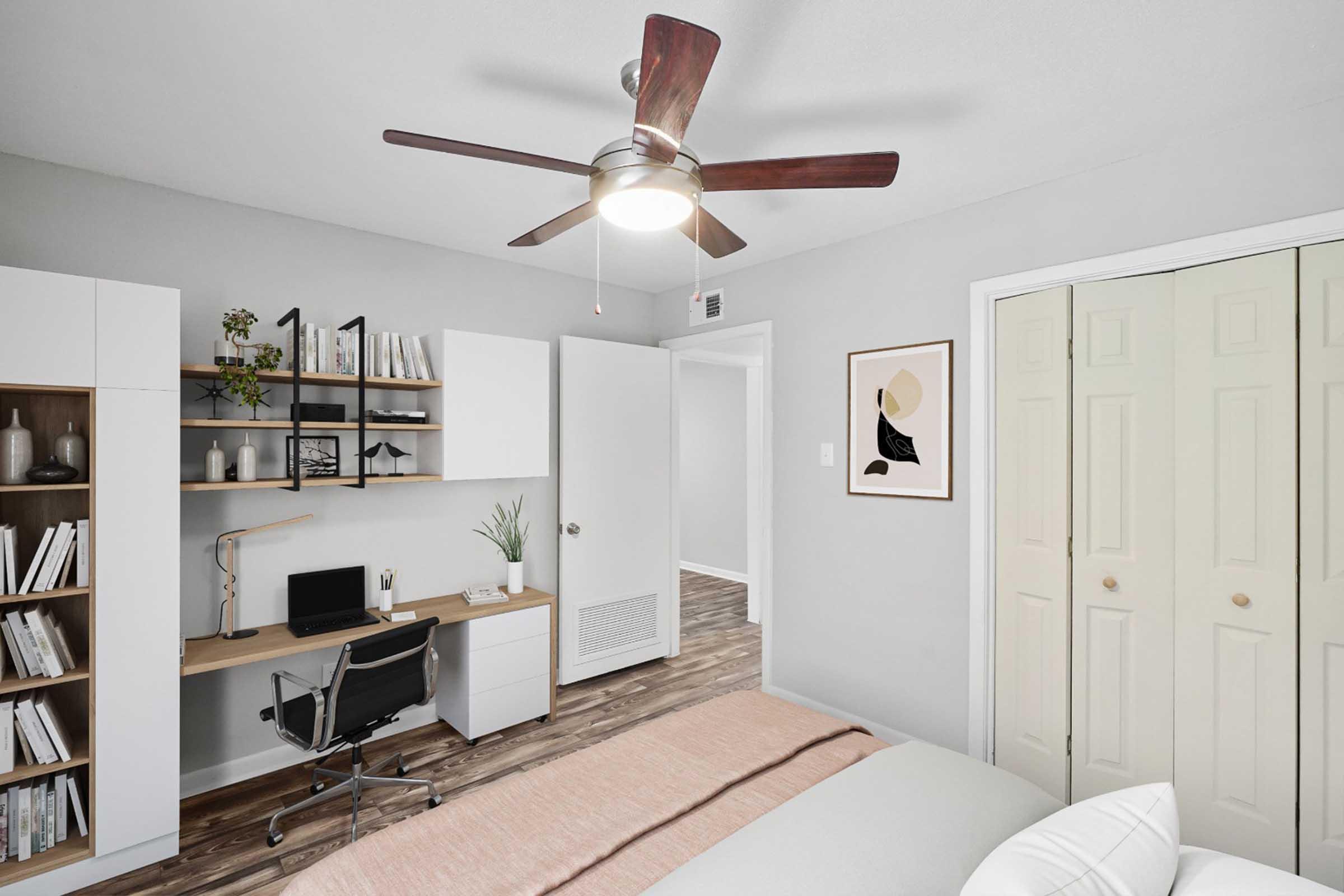 A modern bedroom featuring light gray walls, a ceiling fan, and a cozy bed with a light blanket. There's a small workspace with a desk and chair, along with a bookshelf showcasing decorative items. A closet with double doors is visible, and a framed artwork hangs on the wall.