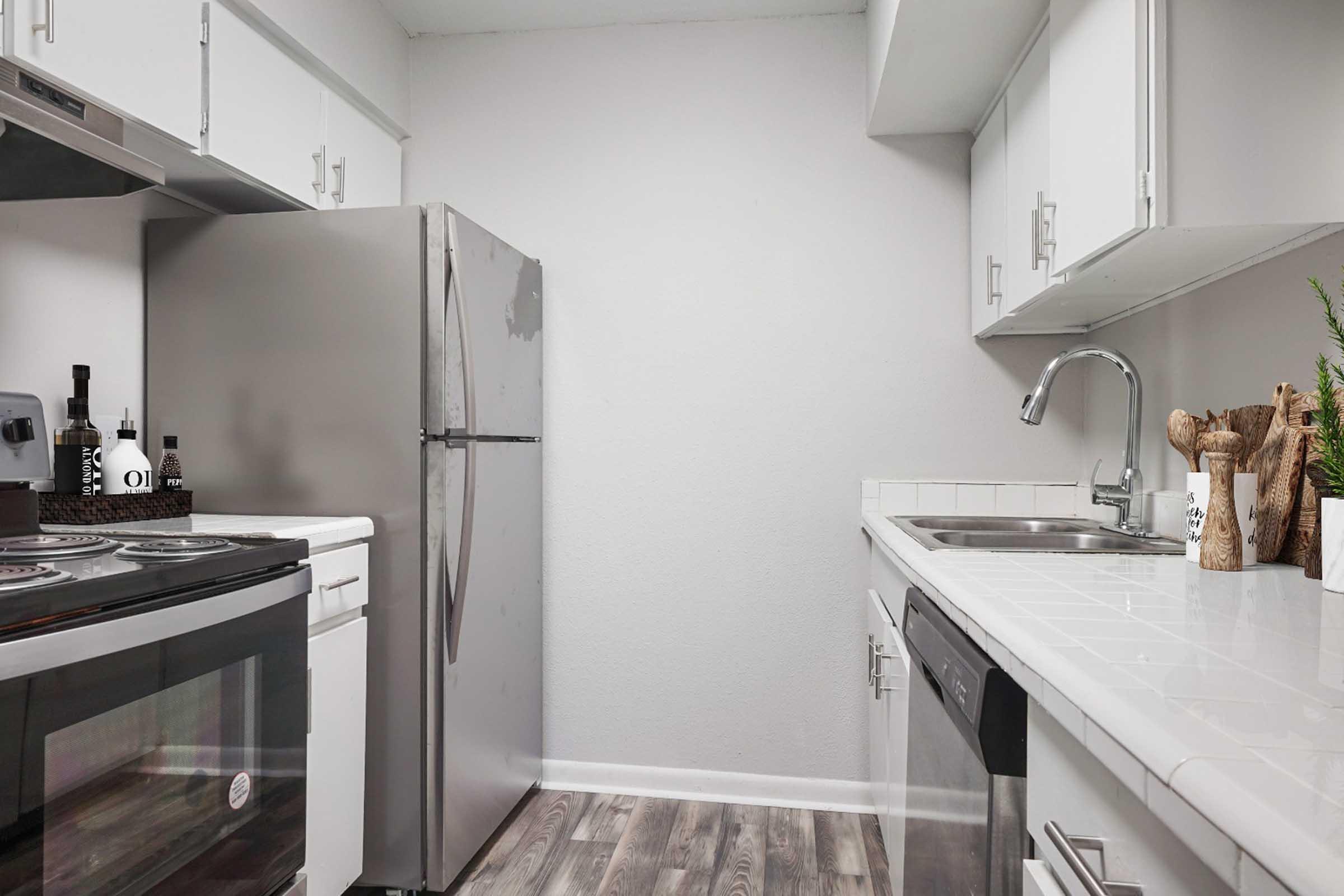 Modern kitchen featuring stainless steel appliances, including a refrigerator and oven. White cabinetry and countertops create a clean look, while a sink is positioned near a window. Decorative elements like kitchen utensils and plants add a touch of warmth to the space.