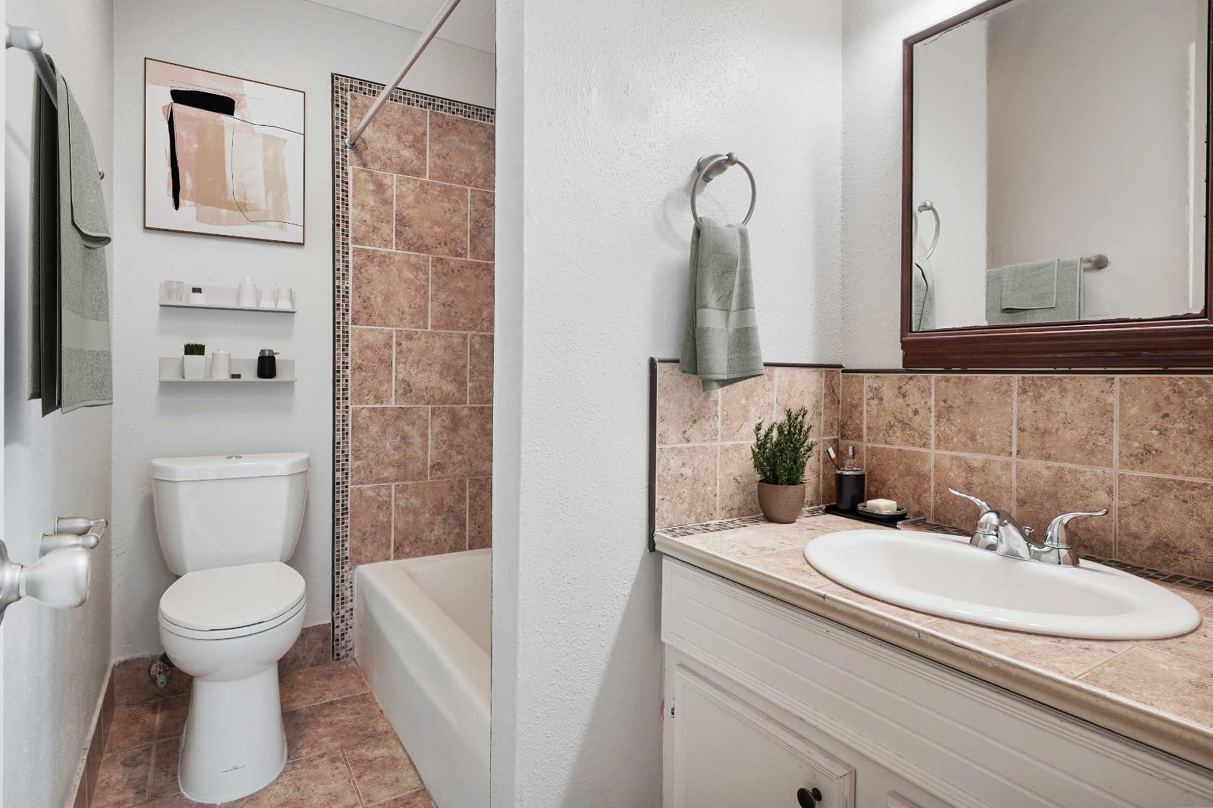 A modern bathroom featuring a toilet, shower, and bathtub. The walls are adorned with beige tiles, and there's a mirror above a sink with a countertop. A small plant decorates the sink area, and there are green towels hanging on the wall. A minimalist shelf holds toiletries.