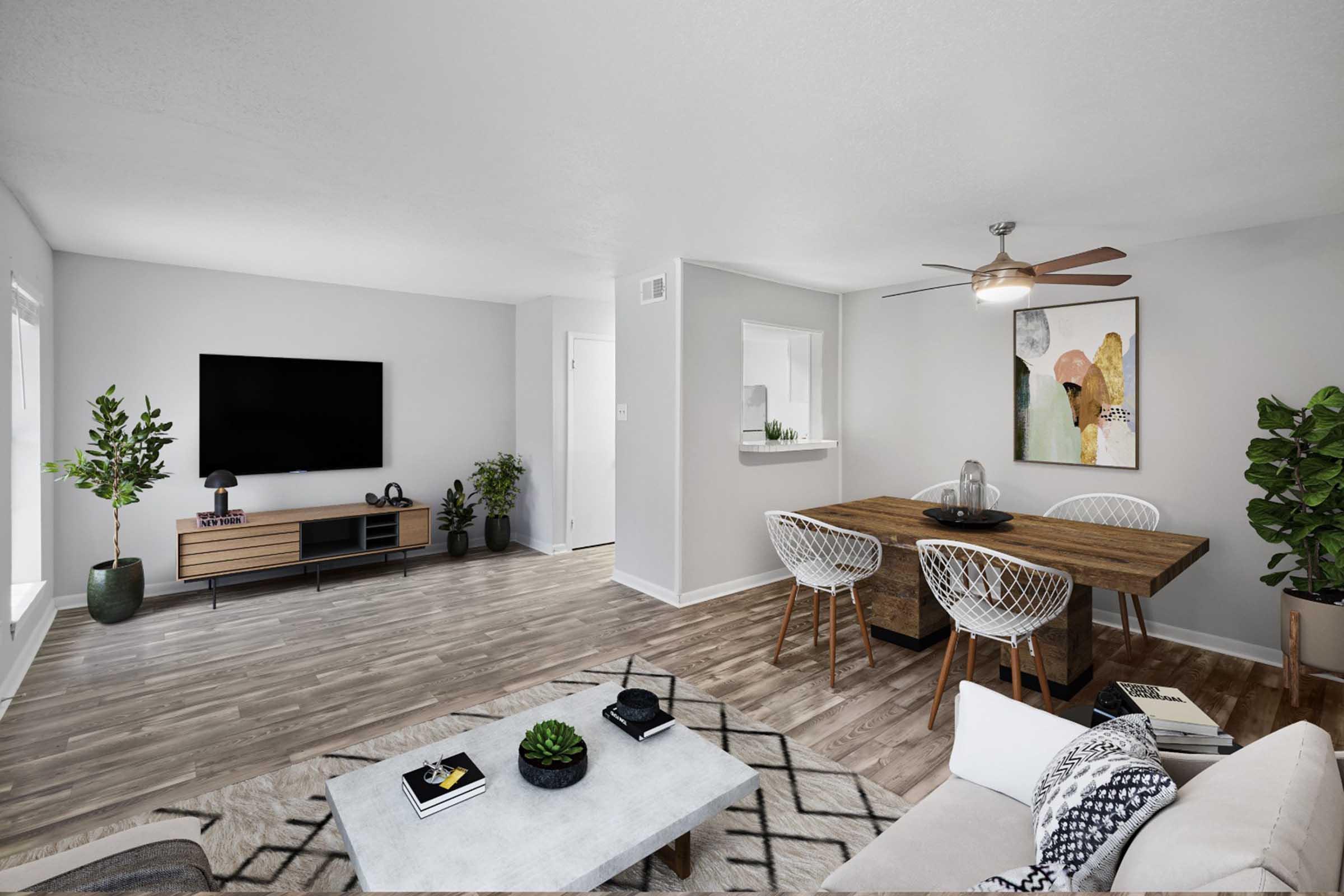 A modern living room with light gray walls and hardwood floors. There is a comfortable sofa with decorative pillows, a coffee table, and a dining area featuring a wooden table and stylish chairs. A large television is mounted on the wall, and indoor plants add greenery to the space. Bright natural light fills the room.