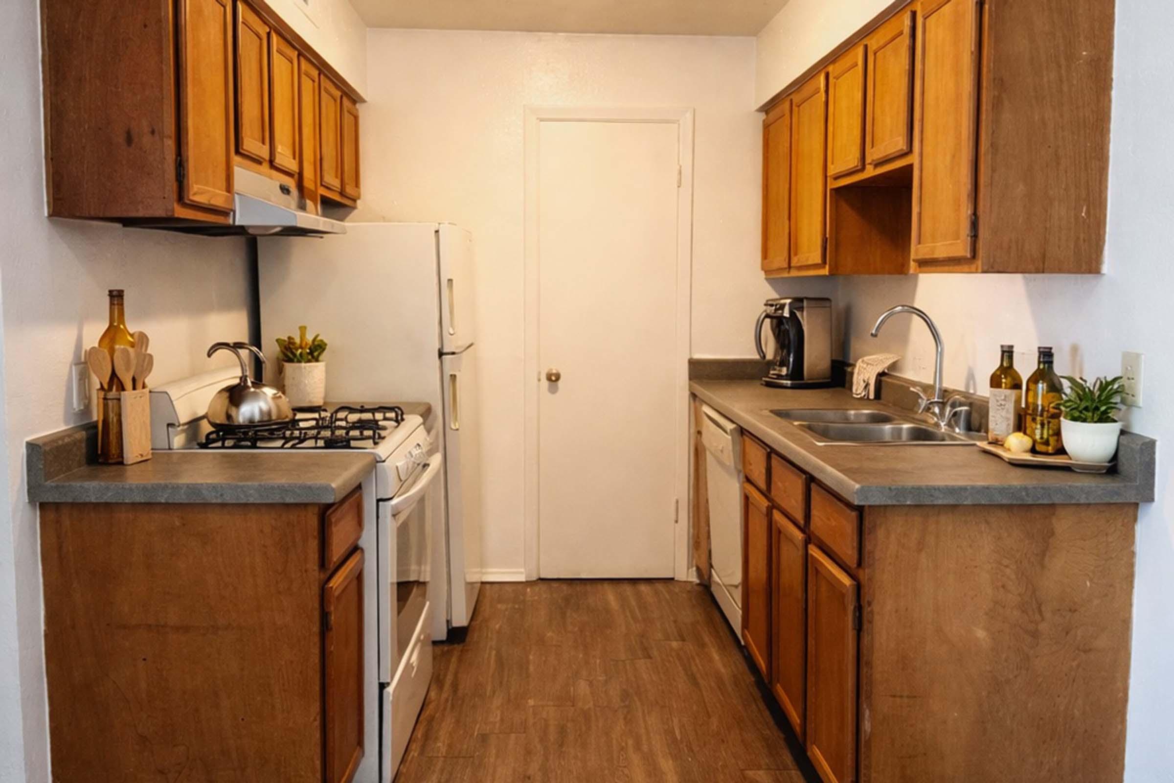 A well-lit kitchen featuring wooden cabinets, a gray countertop, and essential appliances including a white refrigerator, gas stove, and coffee maker. Decorative items include a kettle and several bottles on the counter. A door leads to another room, and there are plants for added decor.