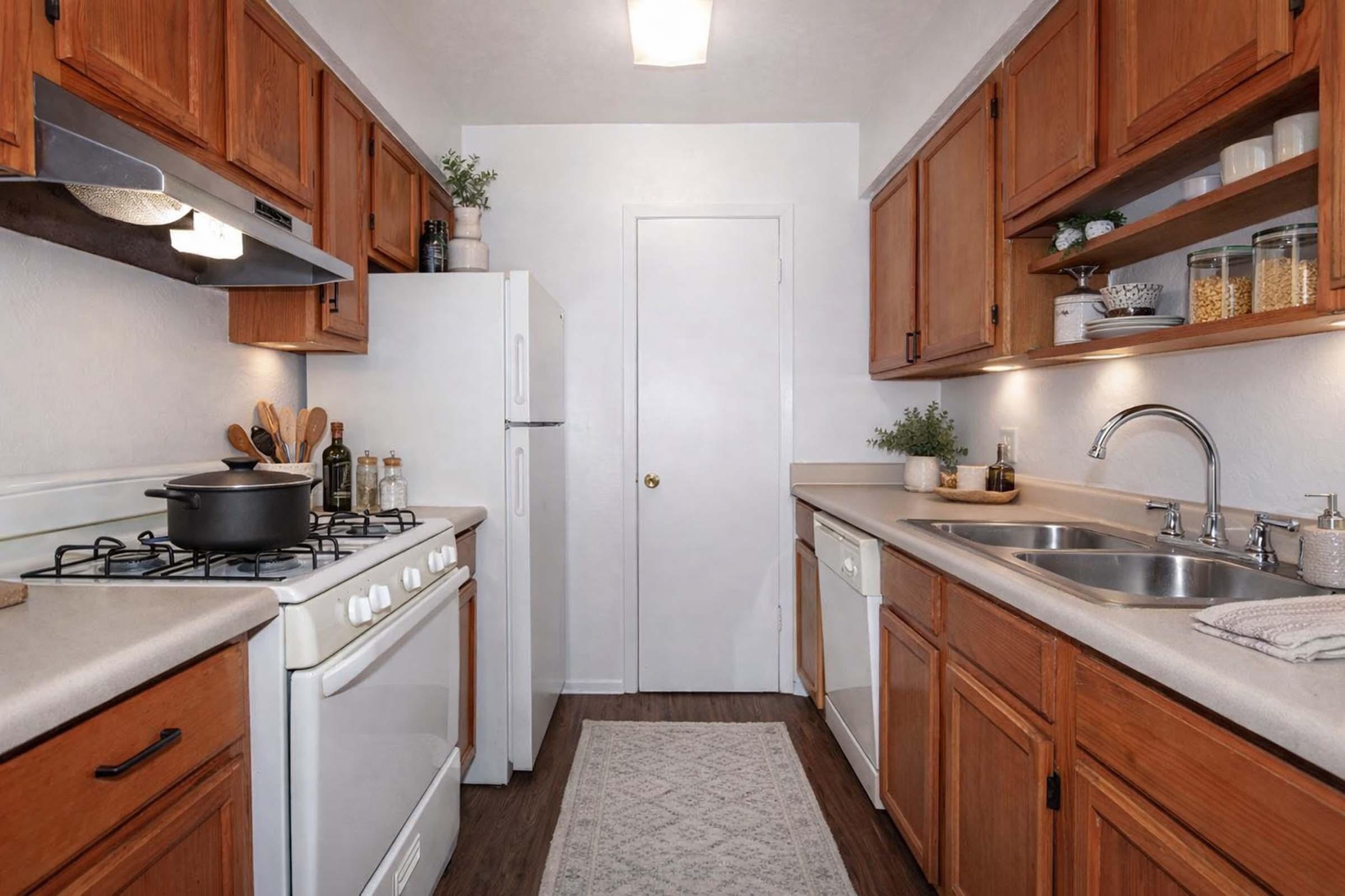 A cozy kitchen featuring wooden cabinets, a gas stove, a white refrigerator, and a double sink. There are decorative plants on the countertop and open shelves displaying jars. The floor is covered with a light rug, and the walls are painted in a neutral color, enhancing the warm and inviting atmosphere.