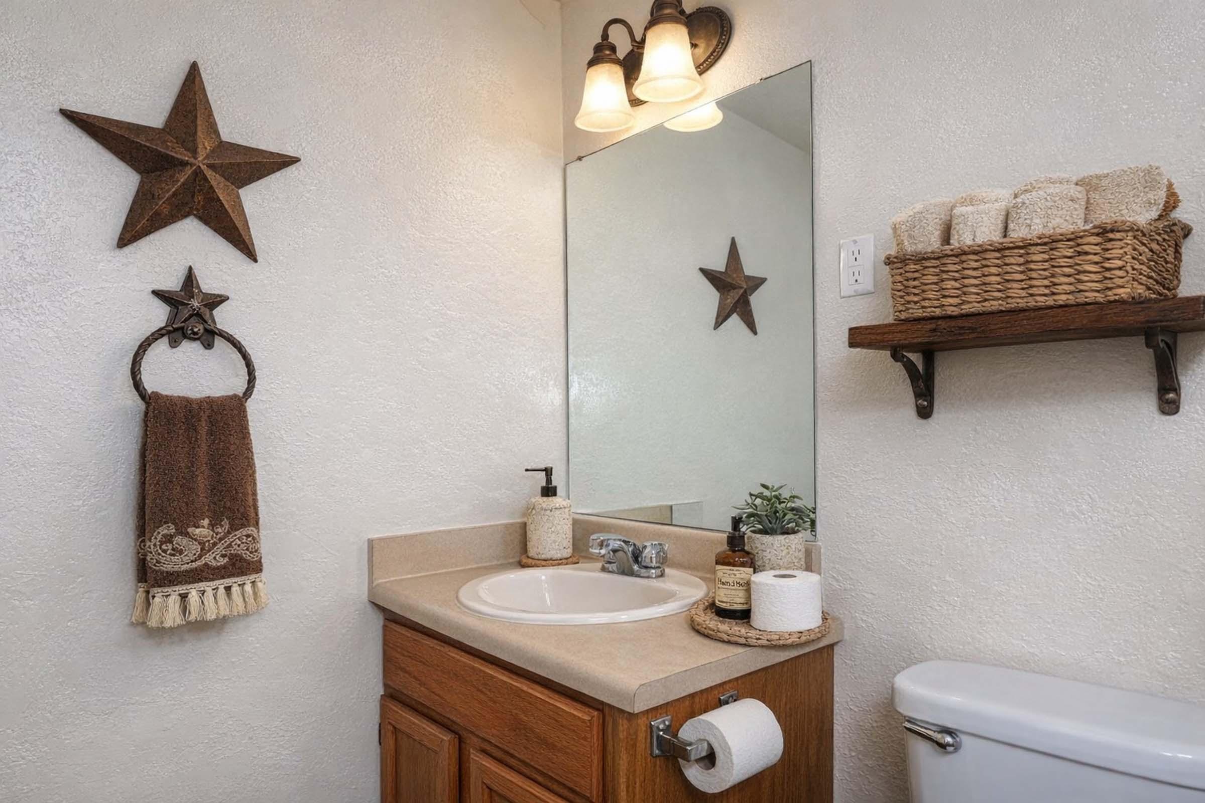 A tidy bathroom featuring a light-colored wall, a wooden vanity with a sink, a round mirror, and a wall-mounted shelf. Decor includes a star motif, a towel rack with a brown towel, and a woven basket of hand towels. The space is well-lit with two light fixtures above the mirror.