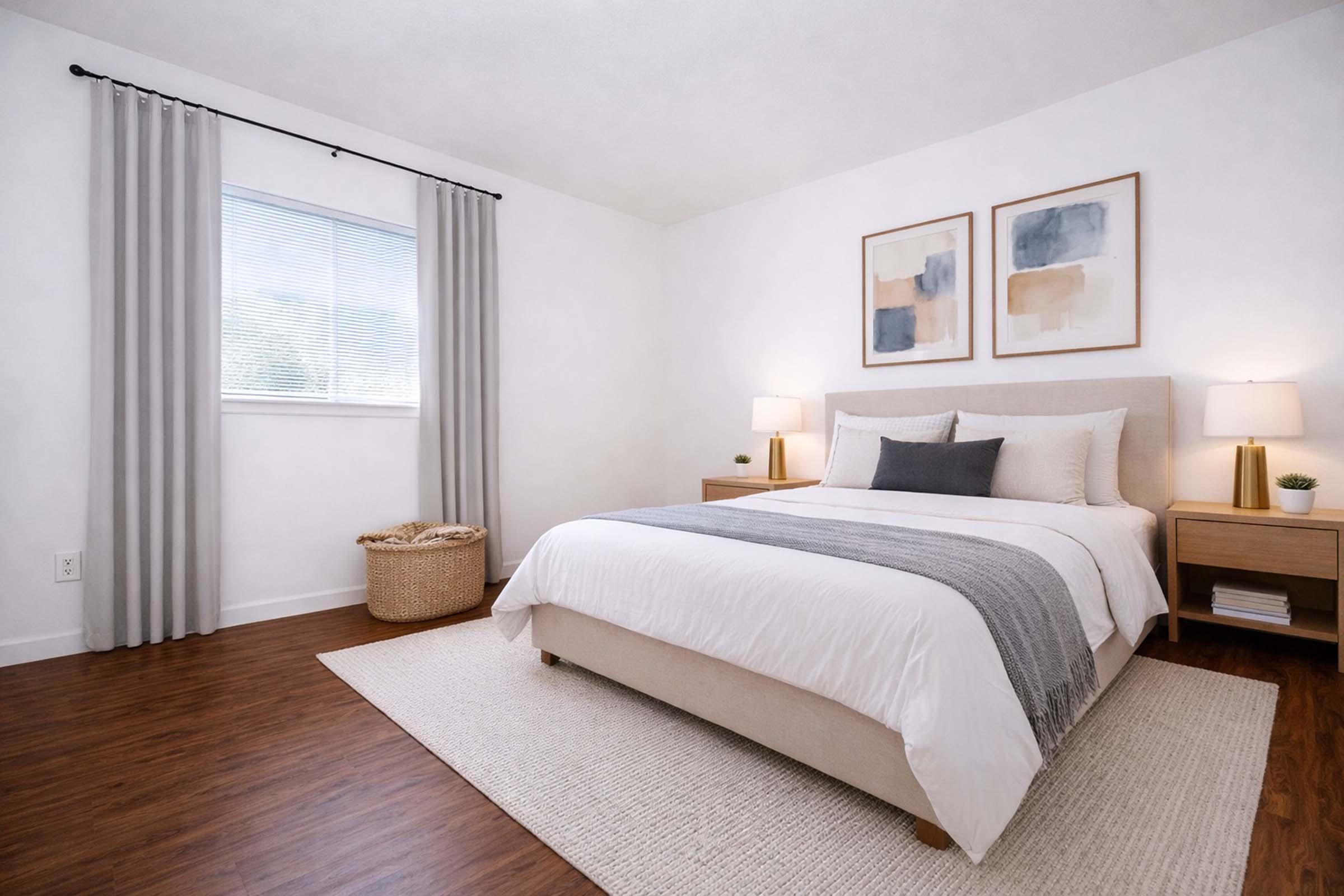 A modern bedroom featuring a neatly made bed with white linens and gray throw. Two light-colored nightstands with lamps flanking the bed. There are abstract art pieces on the wall, a woven basket near the window, and light gray curtains. The floor is hardwood with a light rug.