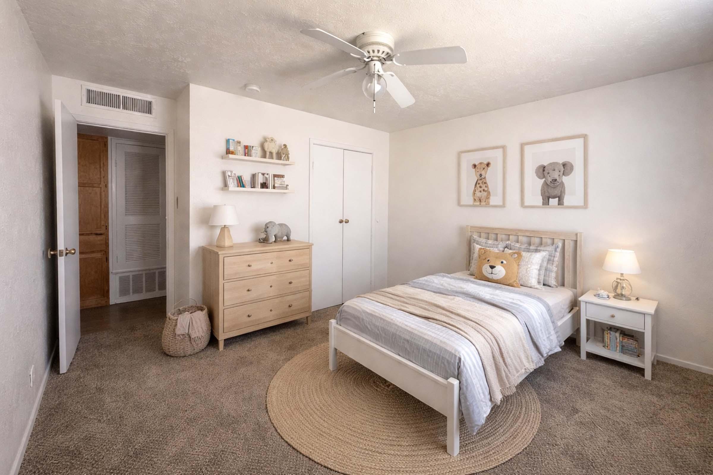 A cozy children's bedroom featuring a white bed with a teddy bear-themed blanket, a dresser, and a soft area rug. The walls are adorned with framed animal prints, and there's a ceiling fan. A small lamp and decorative items on the dresser add warmth to the space.