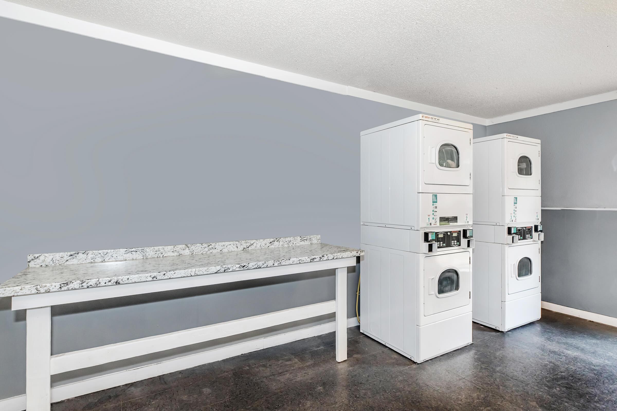 A laundry room featuring a row of stacked white washer and dryer units next to a long granite countertop against a light gray wall. The floor is dark and has a polished appearance.
