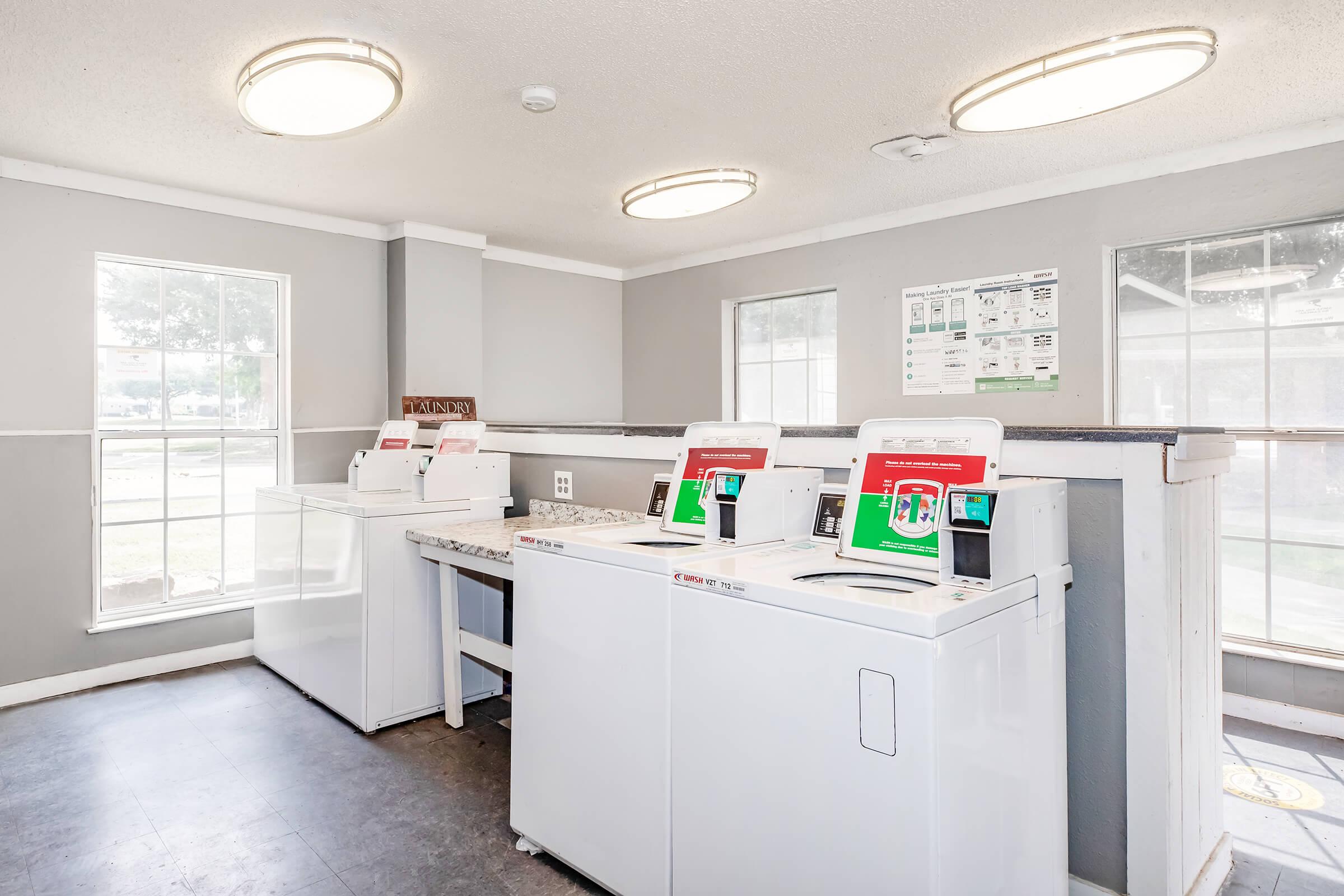 A laundry room featuring several white washing machines and dryers lined up against a gray wall. Large windows allow natural light to enter, and there are informational posters on the wall about laundry operations. The floor is dark, contrasting with the light-colored machines and walls.