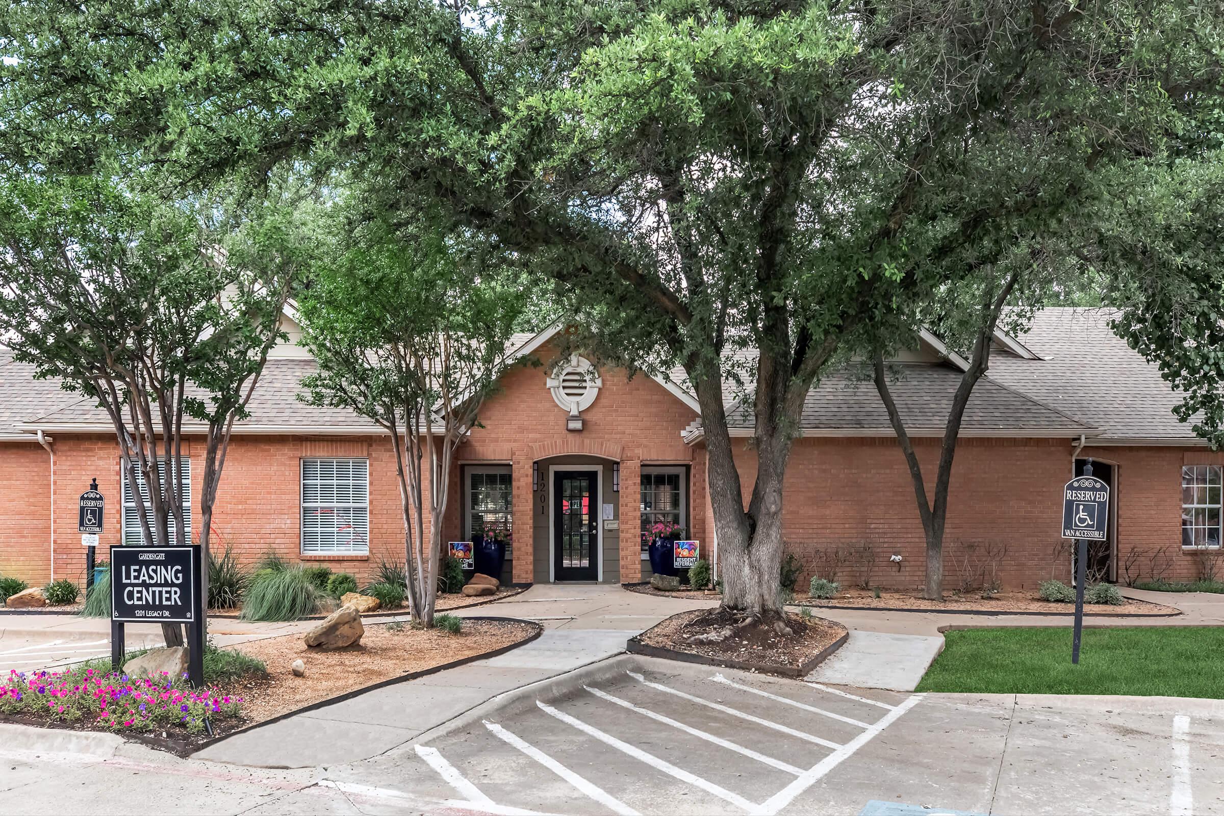 Leasing center of a residential complex with a brick exterior, surrounded by greenery and flower beds. The entrance features double doors and several signs, including one indicating the leasing center. Parking spaces are visible in front of the building.