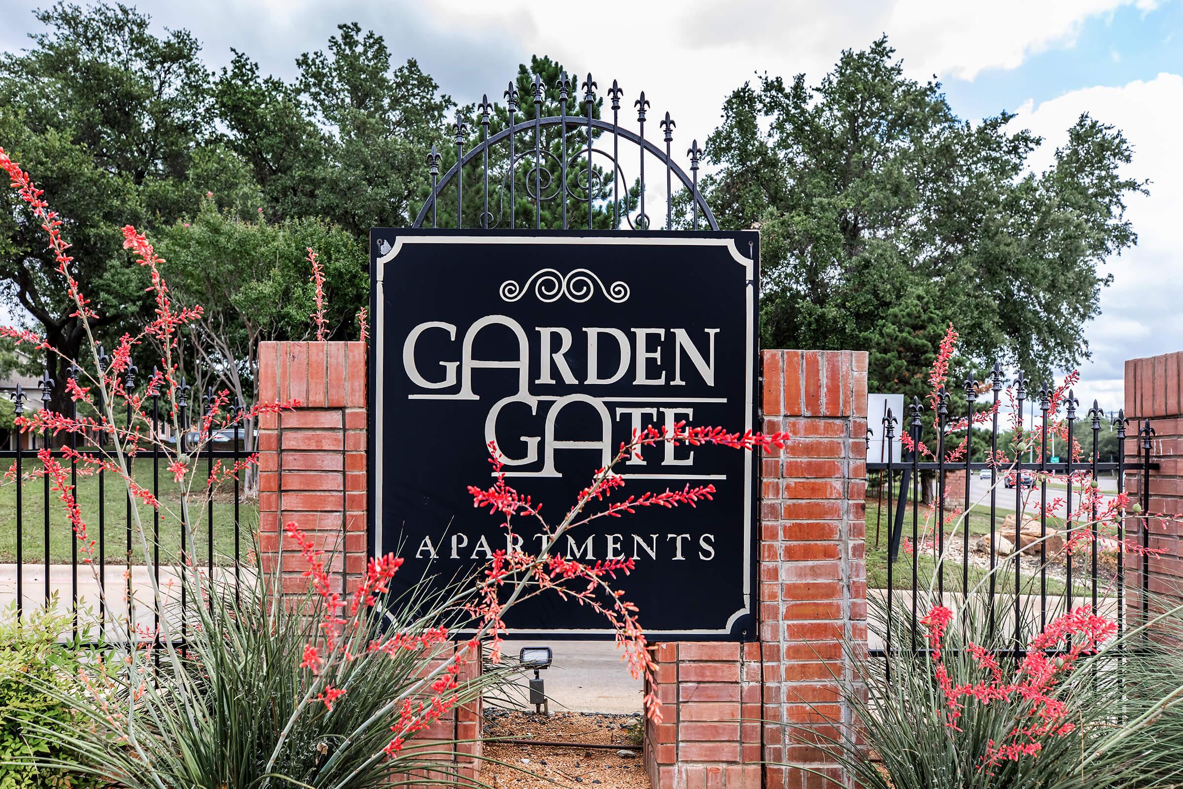 Sign for Garden Gate Apartments, featuring a decorative design in white letters on a dark background. The sign is surrounded by vibrant red flowers and green shrubbery, with trees in the background and a cloudy sky. The overall scene suggests a welcoming residential environment.