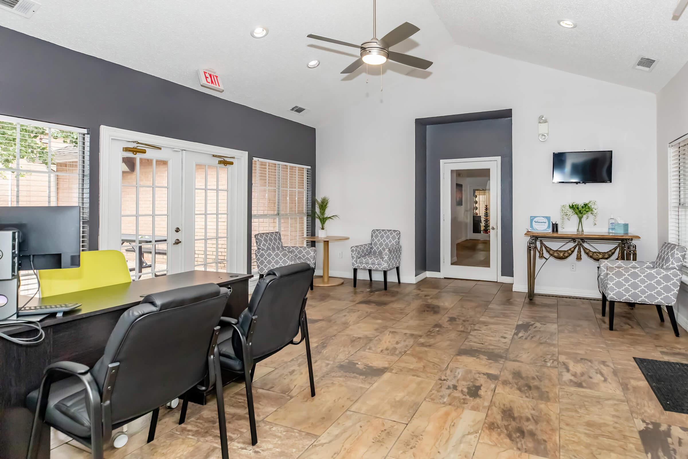 Interior view of a reception area featuring a desk with chairs, a small table, and two upholstered chairs. The space has a cool color palette with gray walls and wooden flooring. Natural light streams in through two sets of doors, and there is a ceiling fan and a wall-mounted TV.