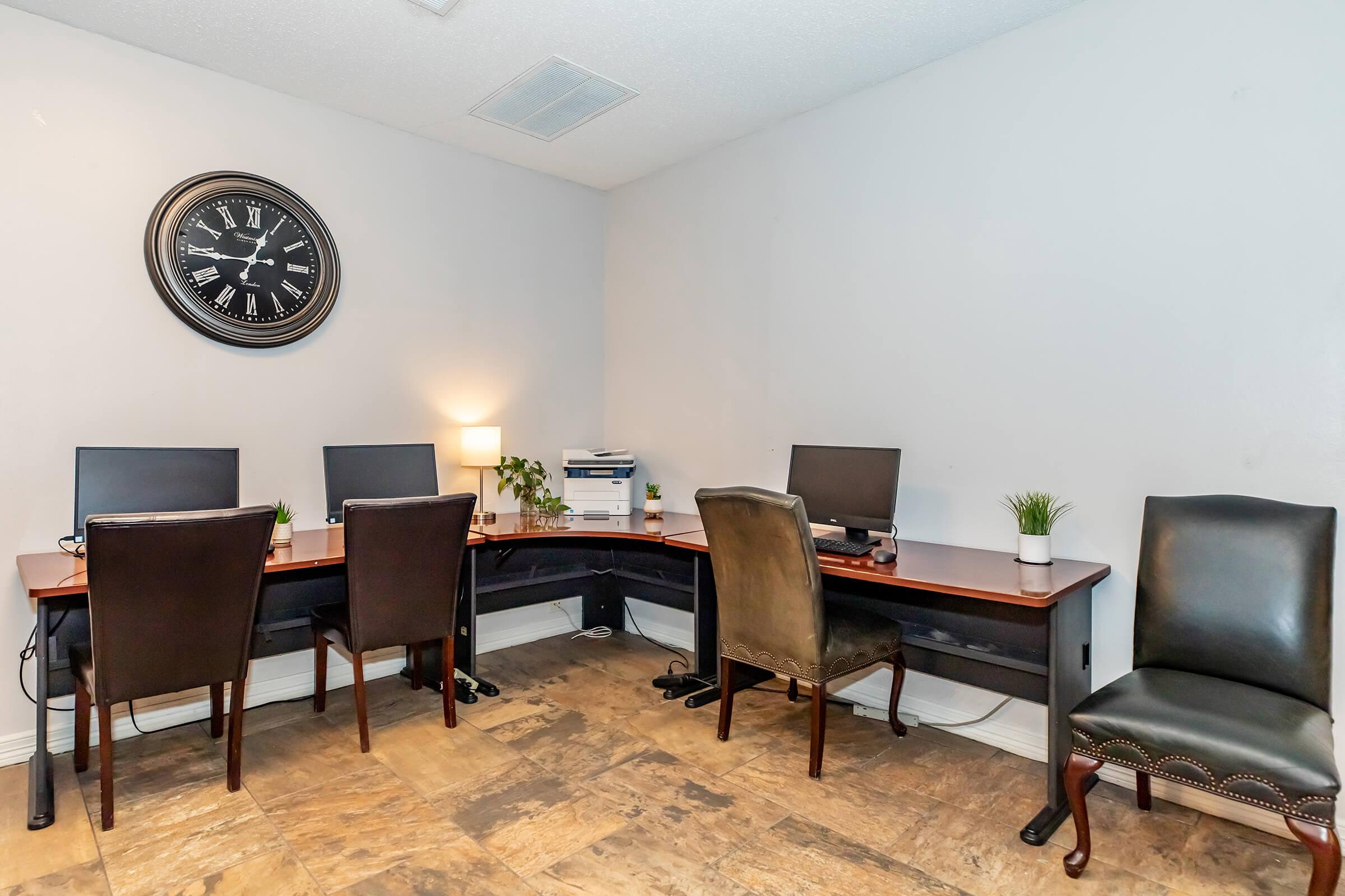 A modern office space featuring four desks with computers, a wall clock, and two types of chairs. The room has a neutral color palette and includes small decorative plants on each desk, with a printer visible in the background. The flooring is tiled, enhancing the professional atmosphere.