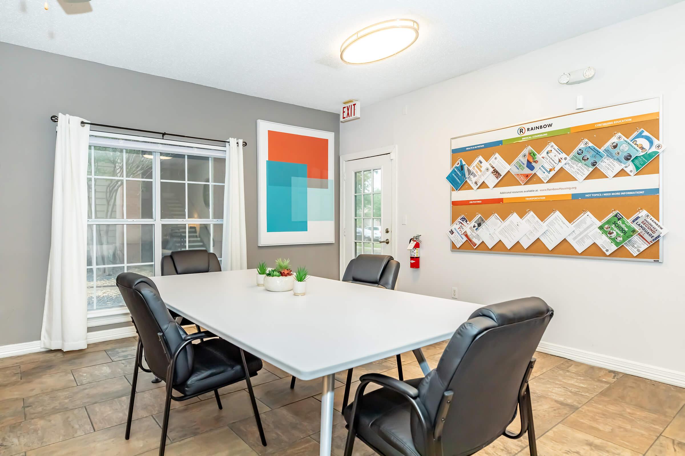 A clean and modern conference room featuring a large white table surrounded by black chairs. A colorful artwork hangs on one wall, while a bulletin board displaying various flyers and information is mounted on another. Natural light streams through a window with white curtains.